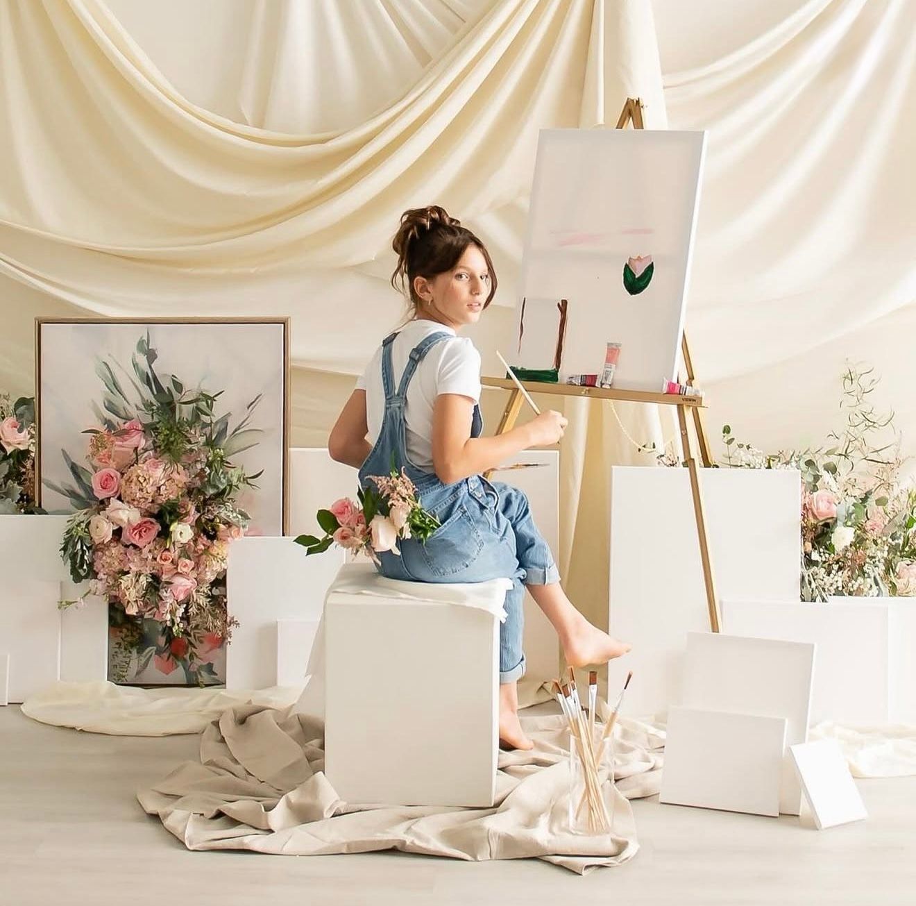 Artist painting on canvas surrounded by floral arrangements in a bright studio with draped fabric backdrop.