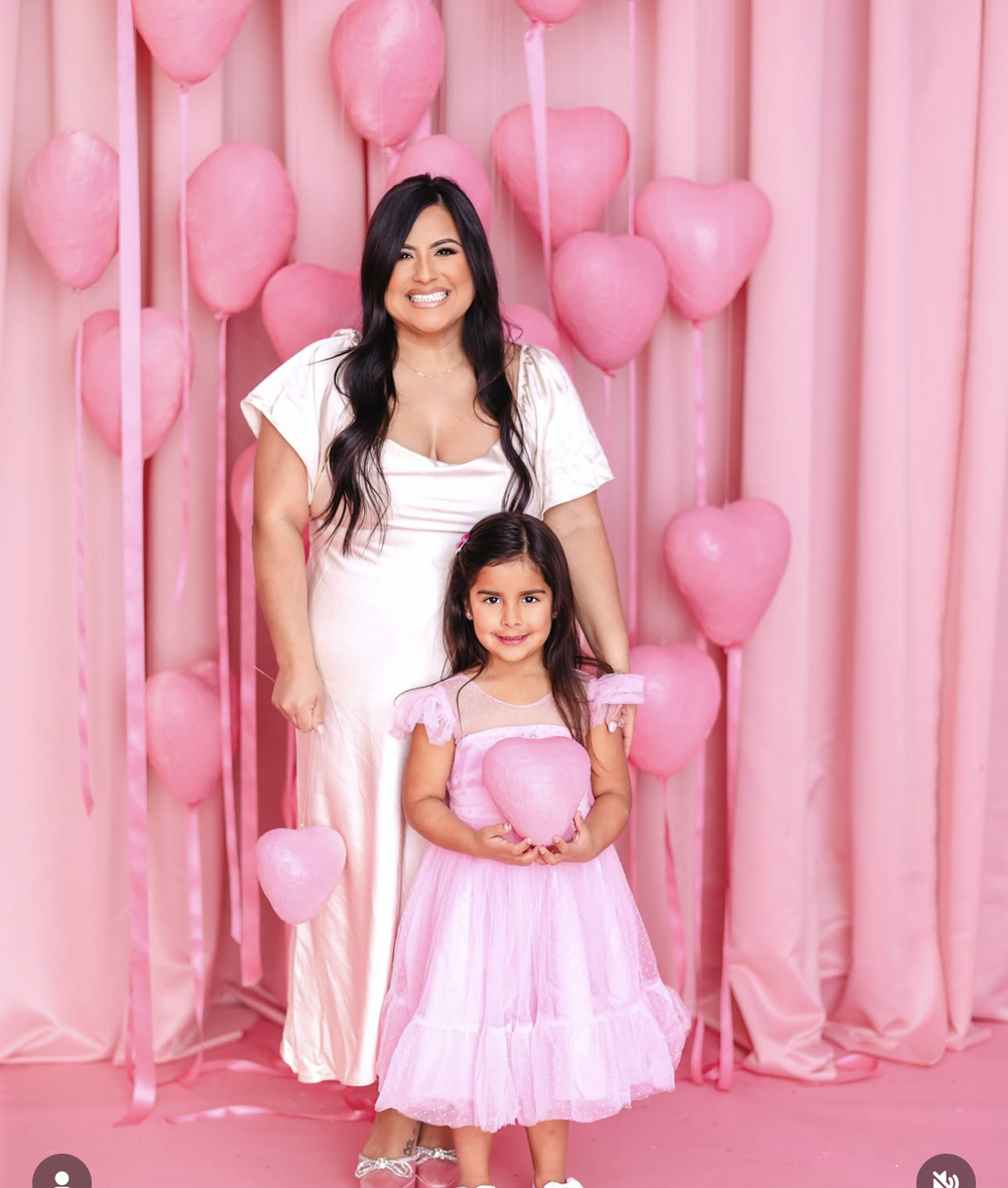 Woman and young girl in pink outfits posing with heart balloons and pink curtain backdrop.