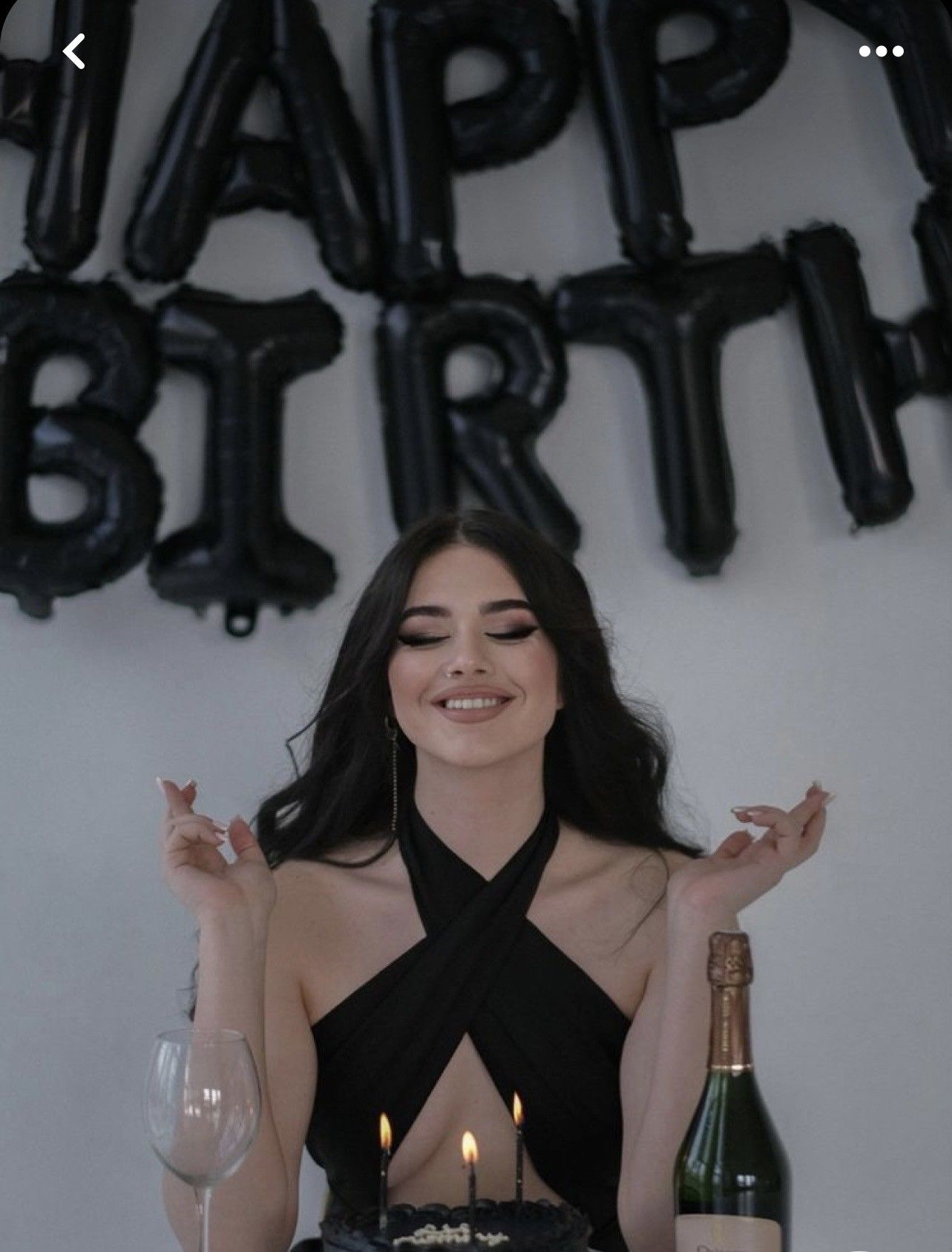 Young woman in black halter dress celebrating birthday with cake, candles, and champagne under black balloon decorations.