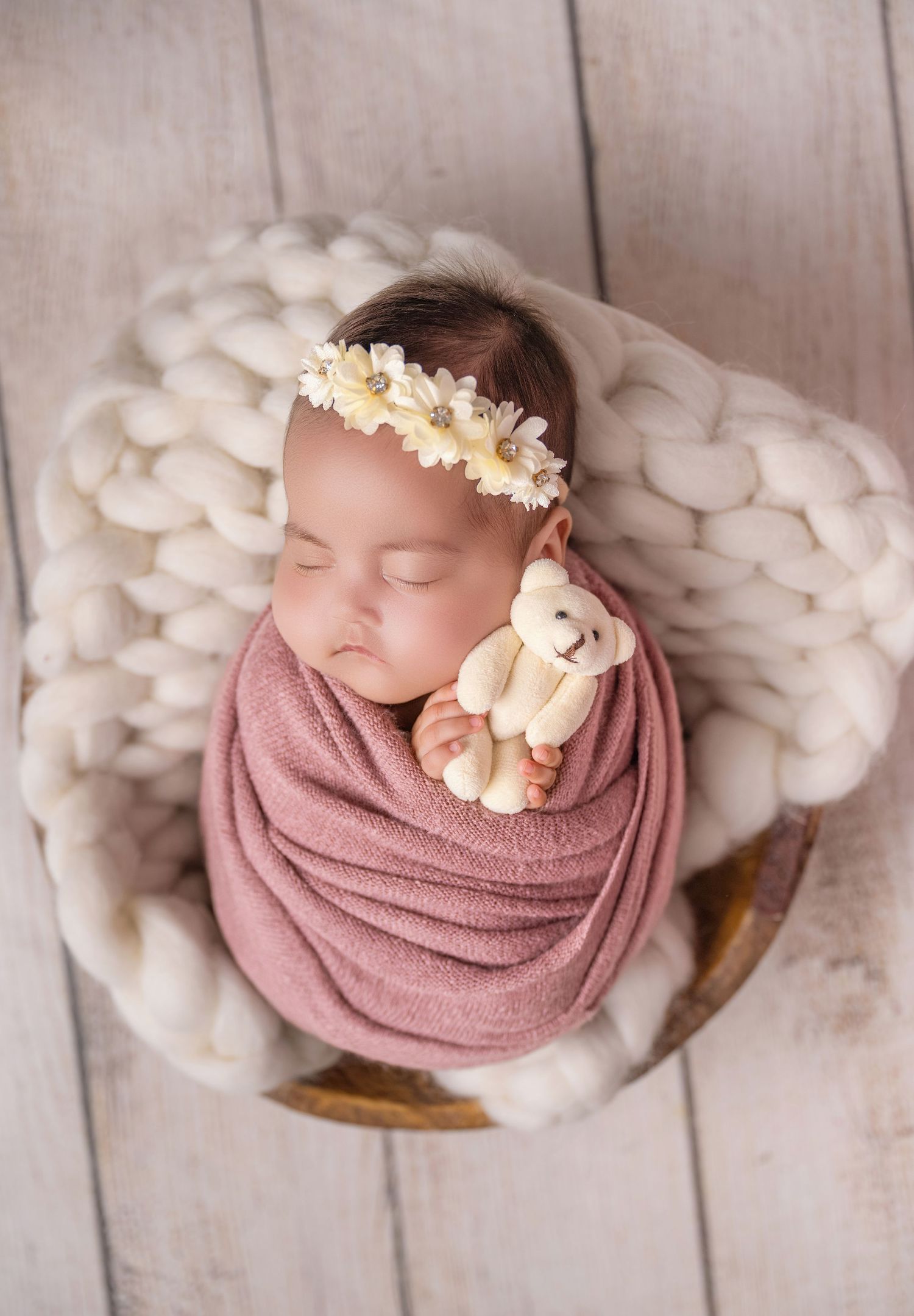 Sleeping newborn baby swaddled in pink wrap, holding a tiny teddy bear, wearing a floral headband in a cozy basket.
