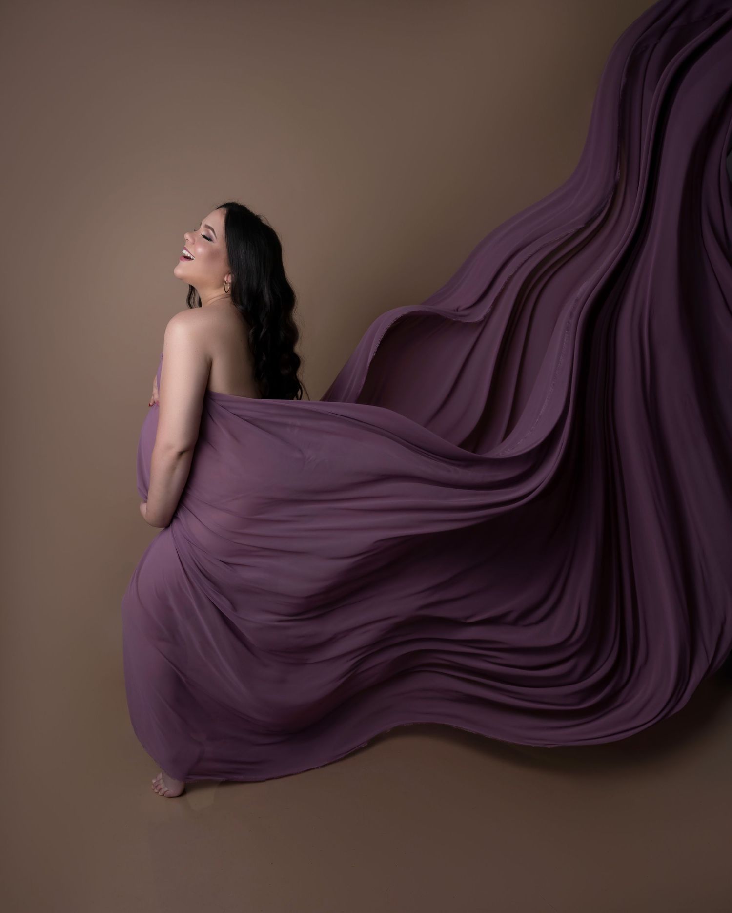 A woman in a flowing purple gown with dramatic fabric billowing behind her against a warm brown background.