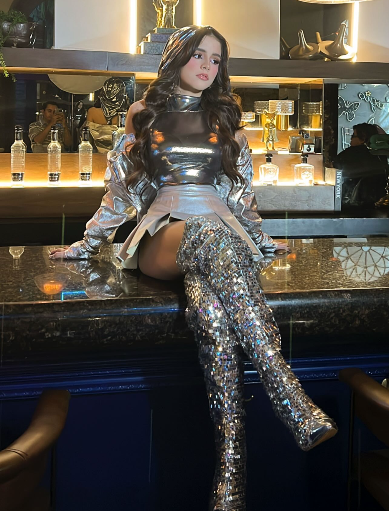 Woman in silver metallic outfit and sequin thigh-high boots posing at a luxury bar.