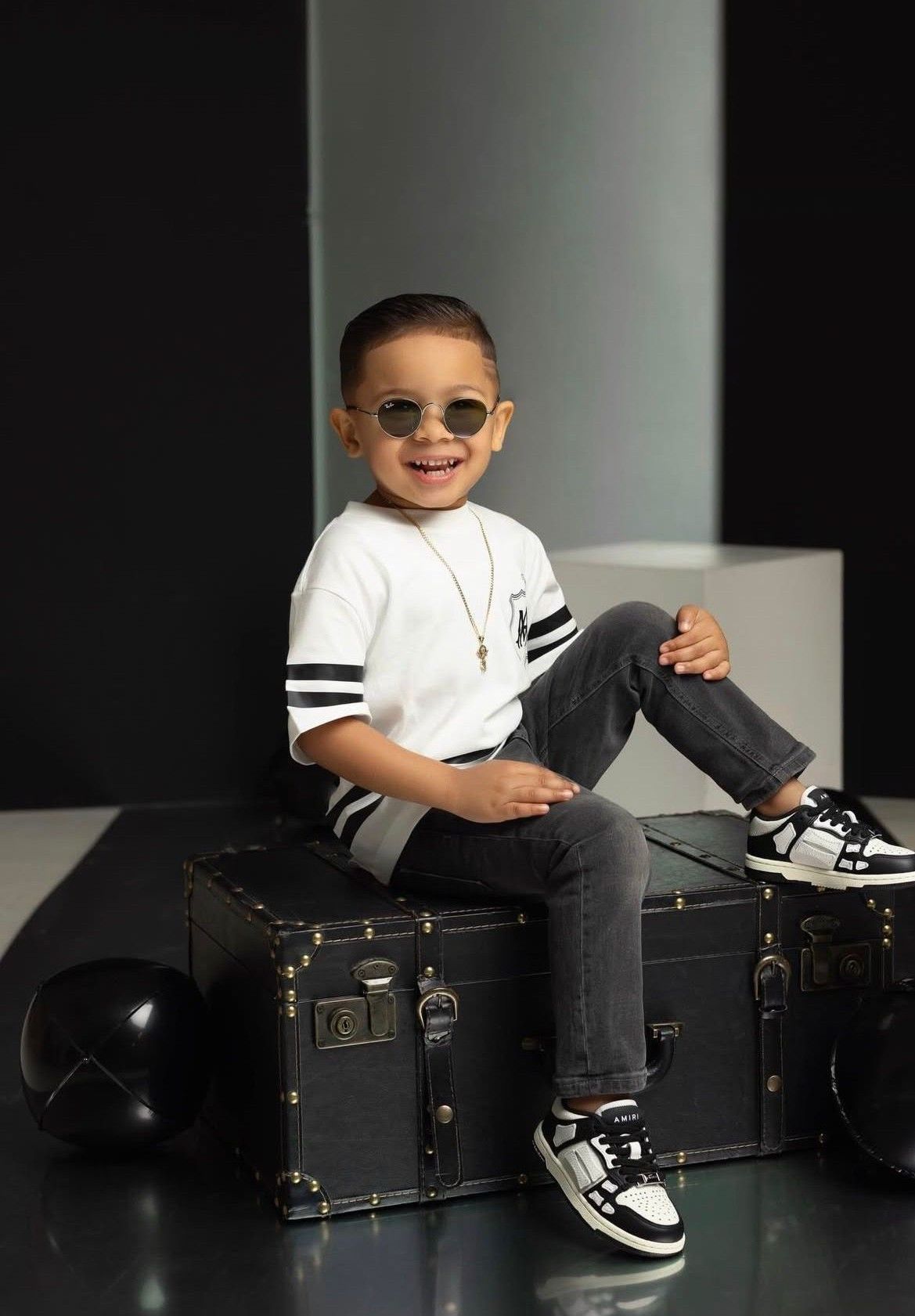 Stylish young boy in sunglasses, white striped tee, gray jeans, sitting on vintage black trunk smiling.