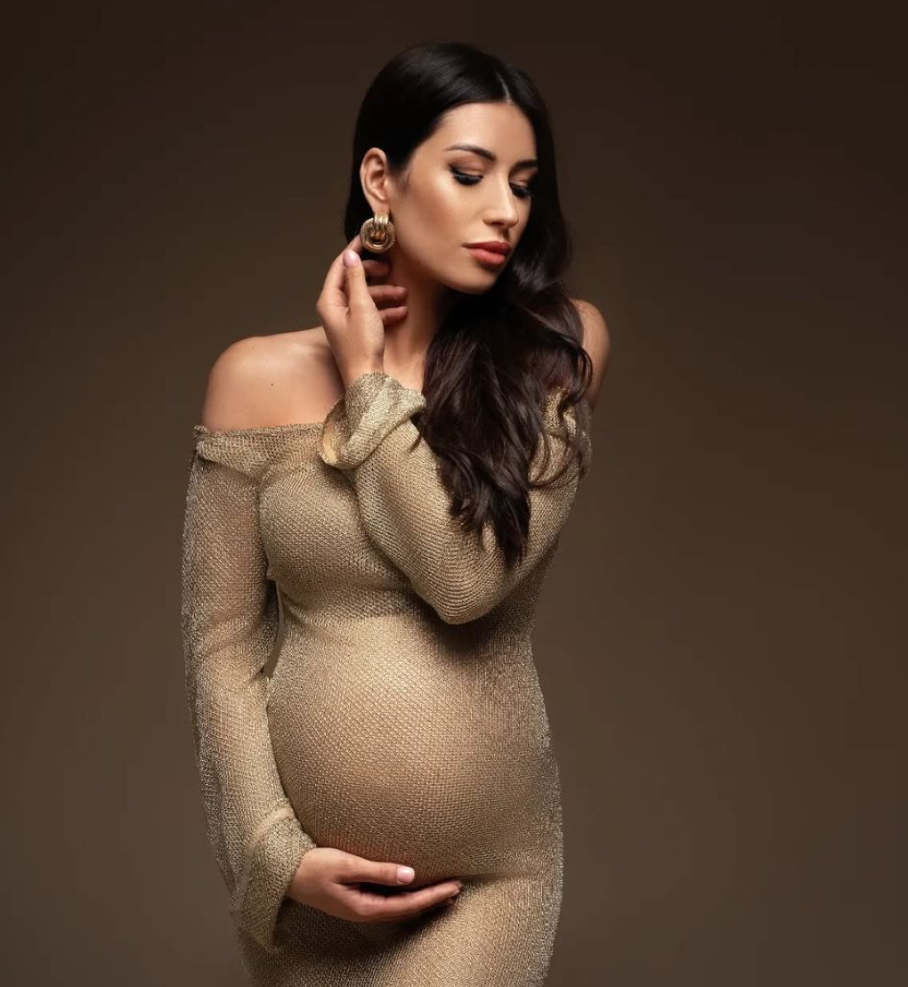 Elegant pregnant woman in a gold off-shoulder gown posing gracefully against a brown background.
