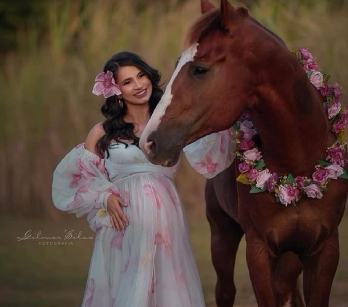 Pregnant woman in floral gown smiling beside a chestnut horse adorned with pink flower garland outdoors.