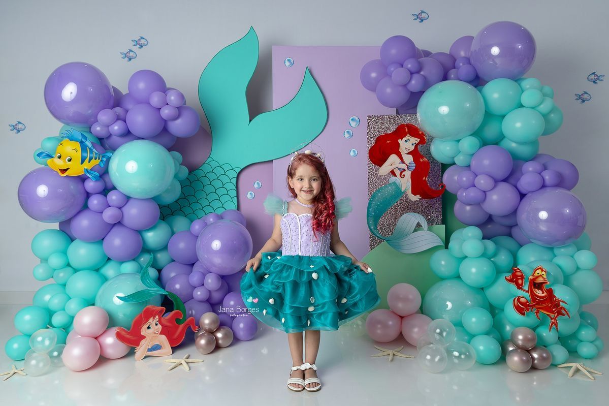 A little girl poses at a Little Mermaid themed birthday party with purple and teal balloon decorations.