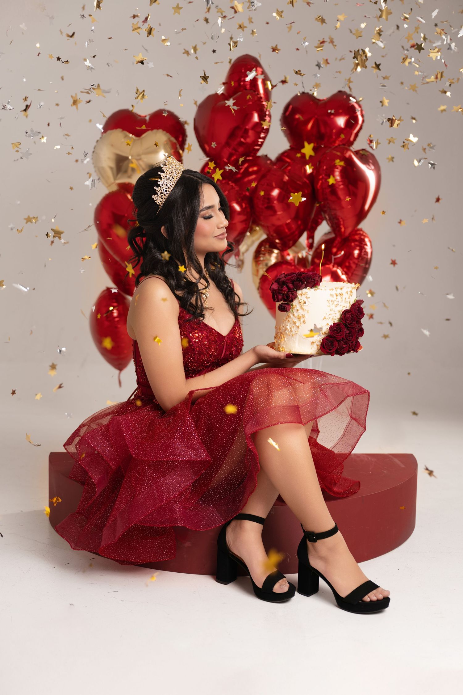Young woman in red sequin dress and crown holds cake, surrounded by heart balloons and falling gold confetti.