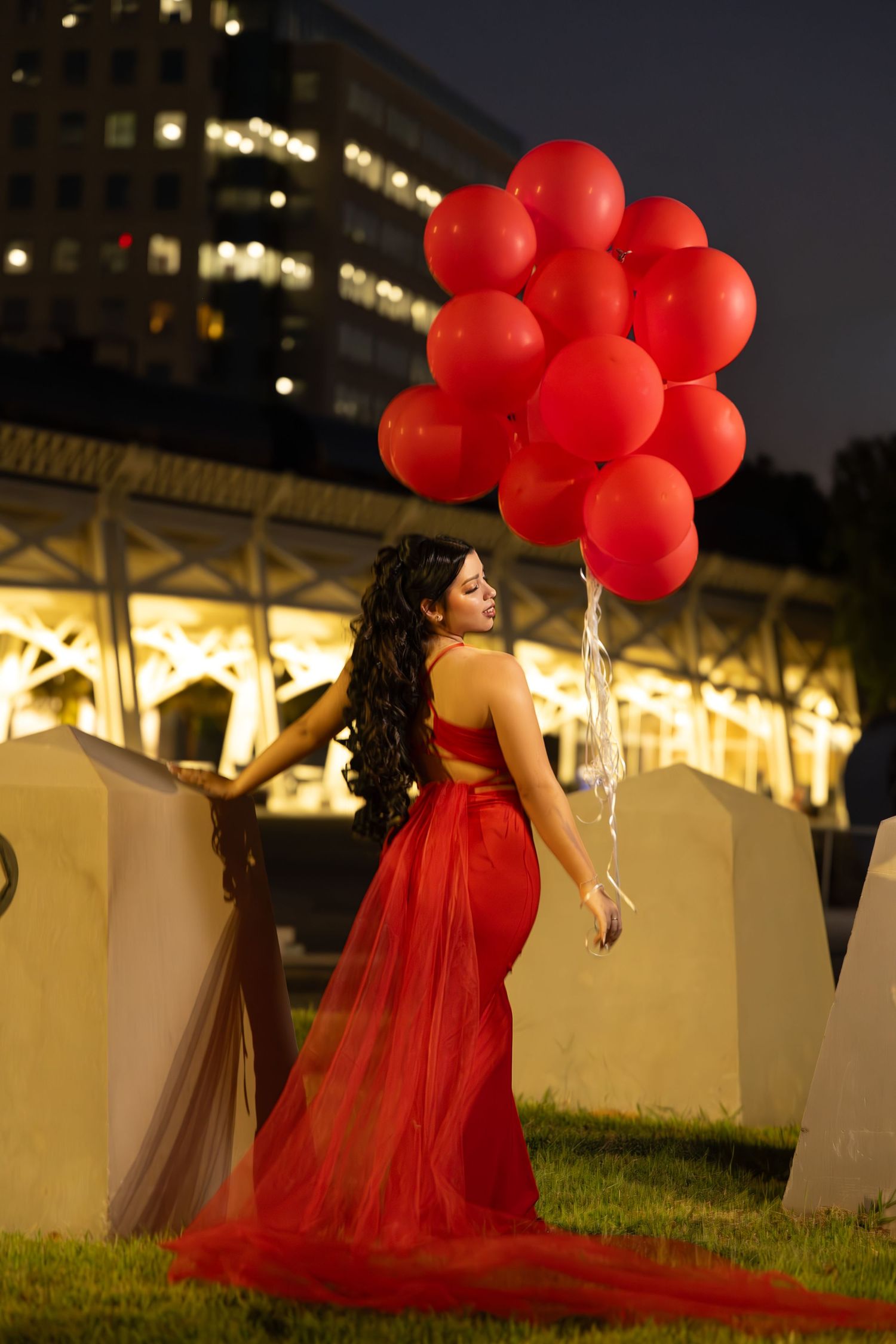 Woman in red gown holding large bunch of red balloons outdoors at night with illuminated buildings behind her.