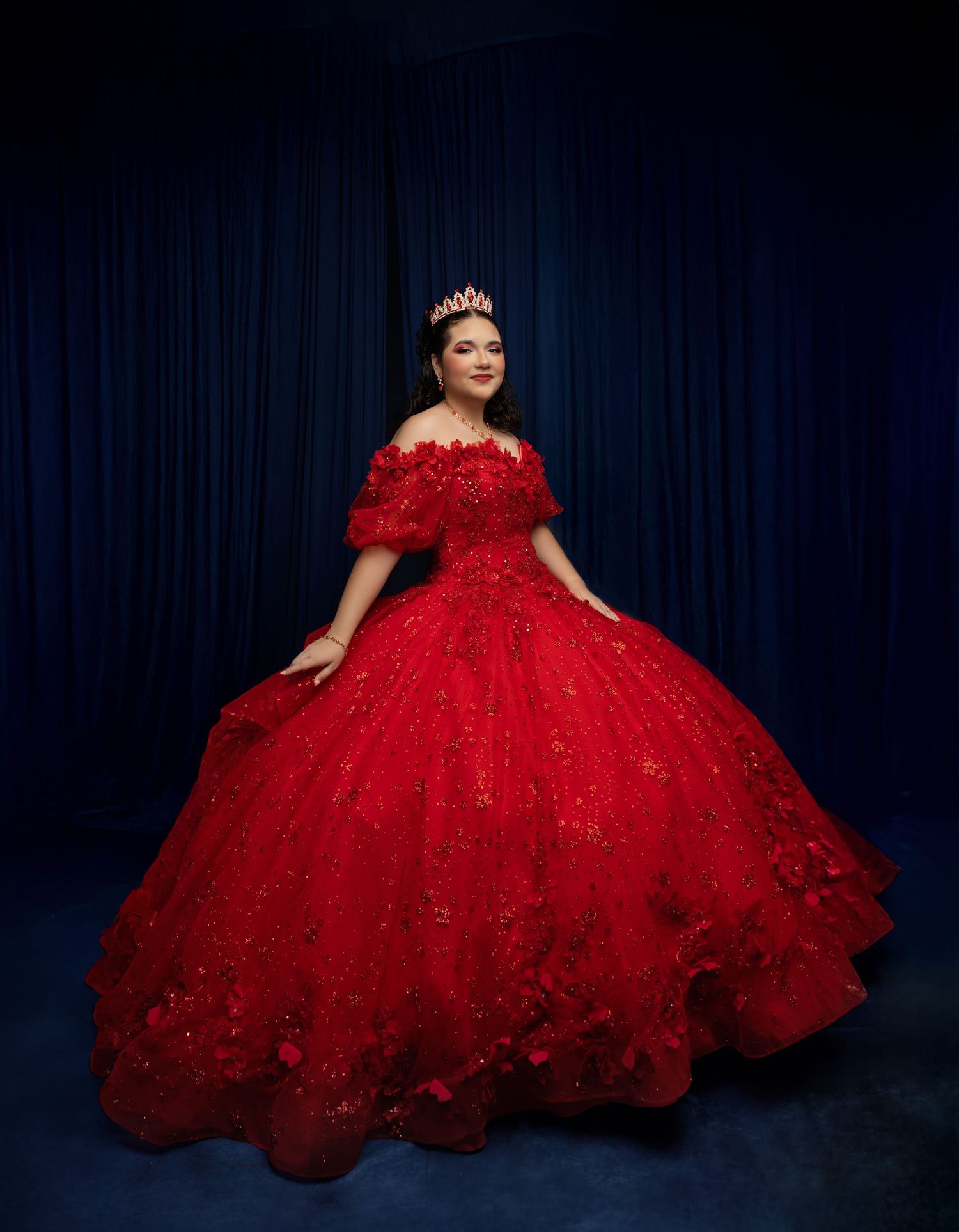 Young woman in a stunning red ball gown with floral details and tiara poses elegantly for a quinceañera portrait.