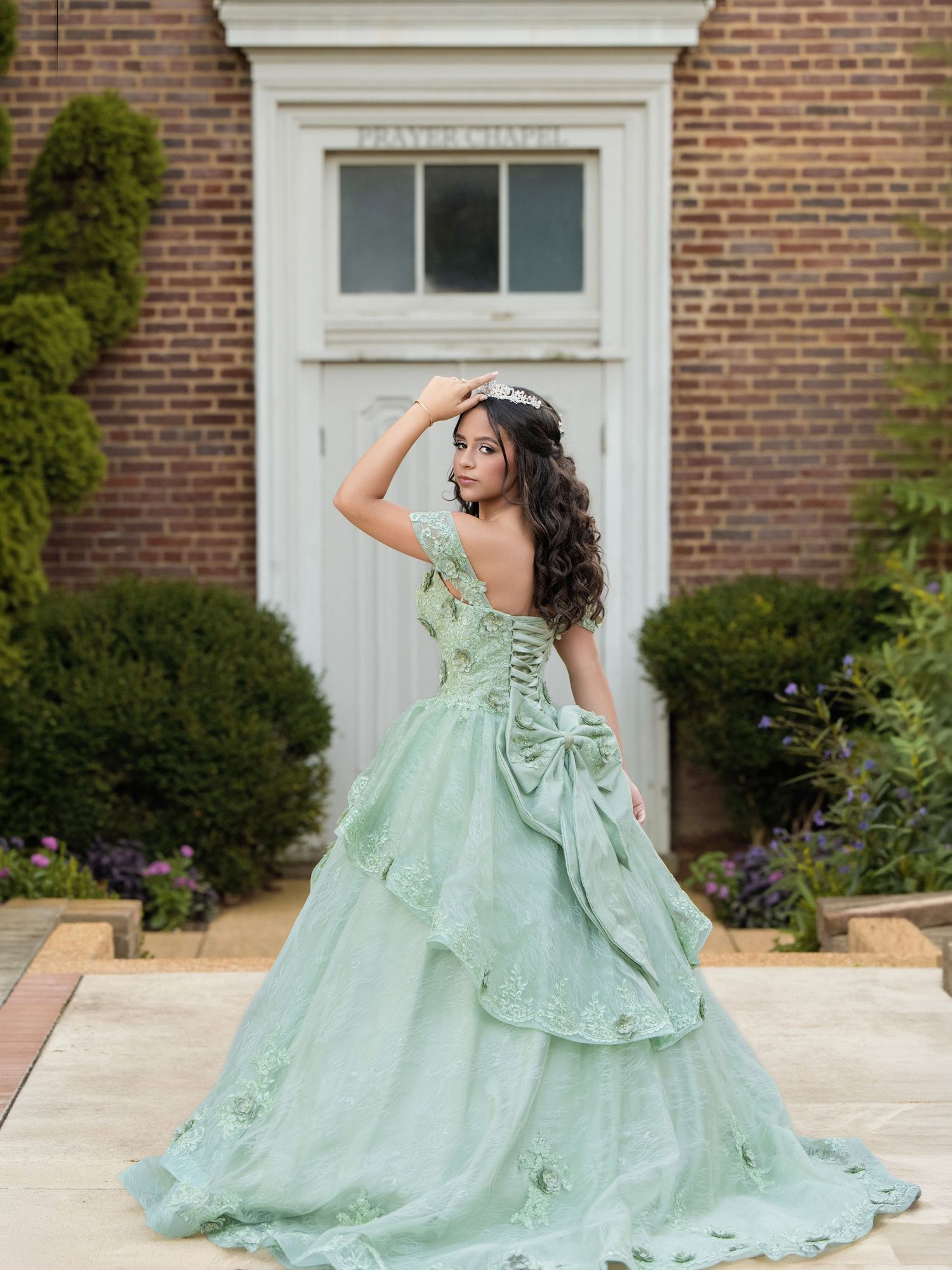 Young woman in elegant mint green quinceañera ball gown with lace details and bow, posing outdoors near brick building.