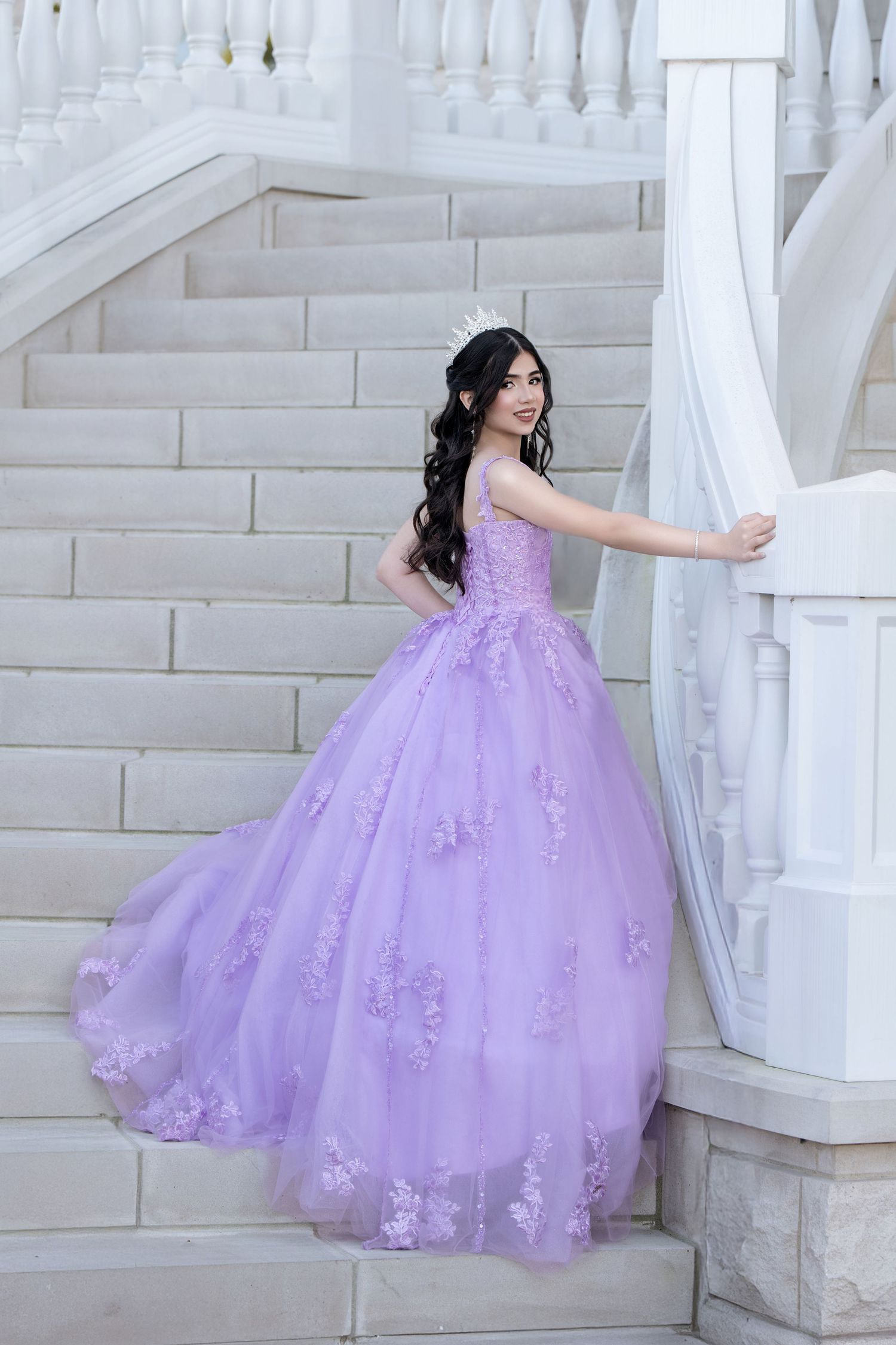 Young woman in lavender ball gown with floral appliques and tiara poses gracefully on white marble staircase.