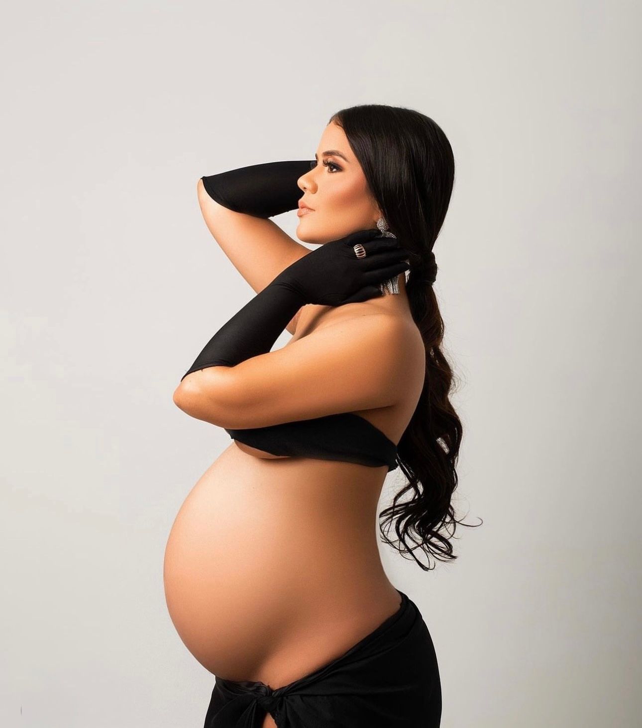 Pregnant woman in black gloves and skirt posing sideways, showcasing baby bump in a professional maternity photo shoot.