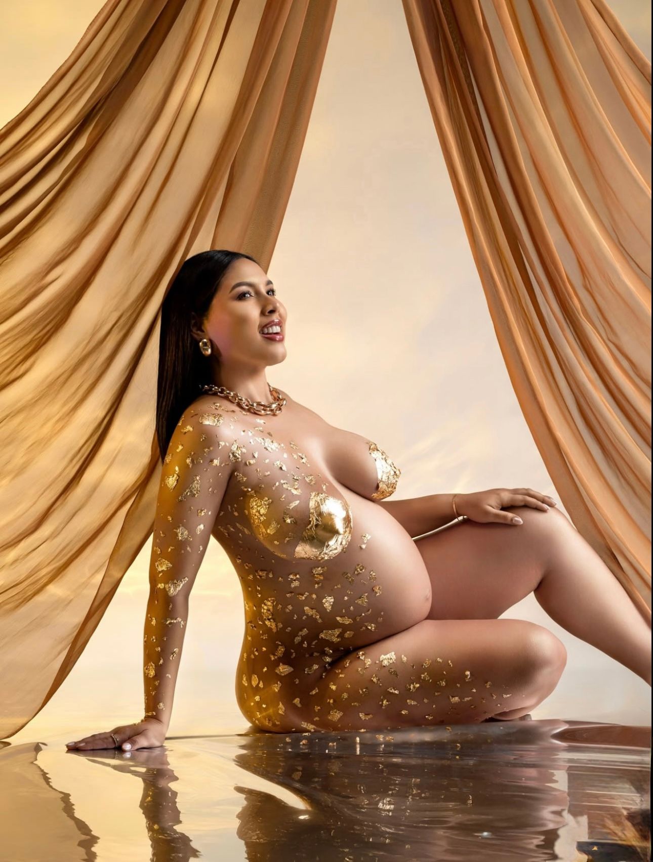 Pregnant woman posed elegantly in golden body paint with draped fabric backdrop in a glamorous maternity photo shoot.