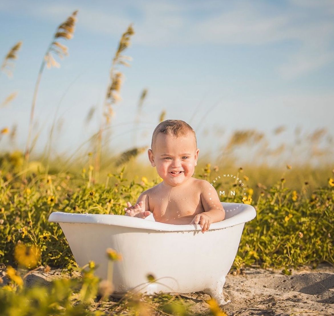 Smiling baby sitting in a white clawfoot bathtub outdoors on a sandy beach surrounded by sea oats and yellow wildflowers.