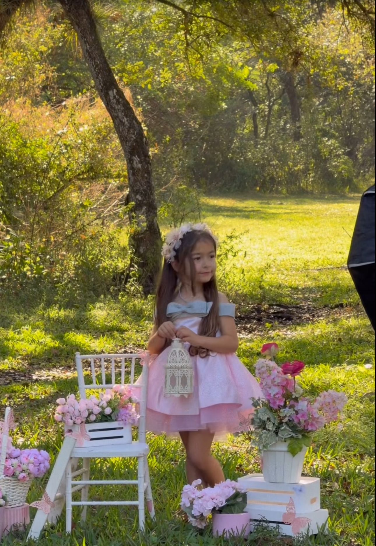 Young girl in pink tutu dress holds birdcage among floral decorations outdoors during golden hour photo shoot.