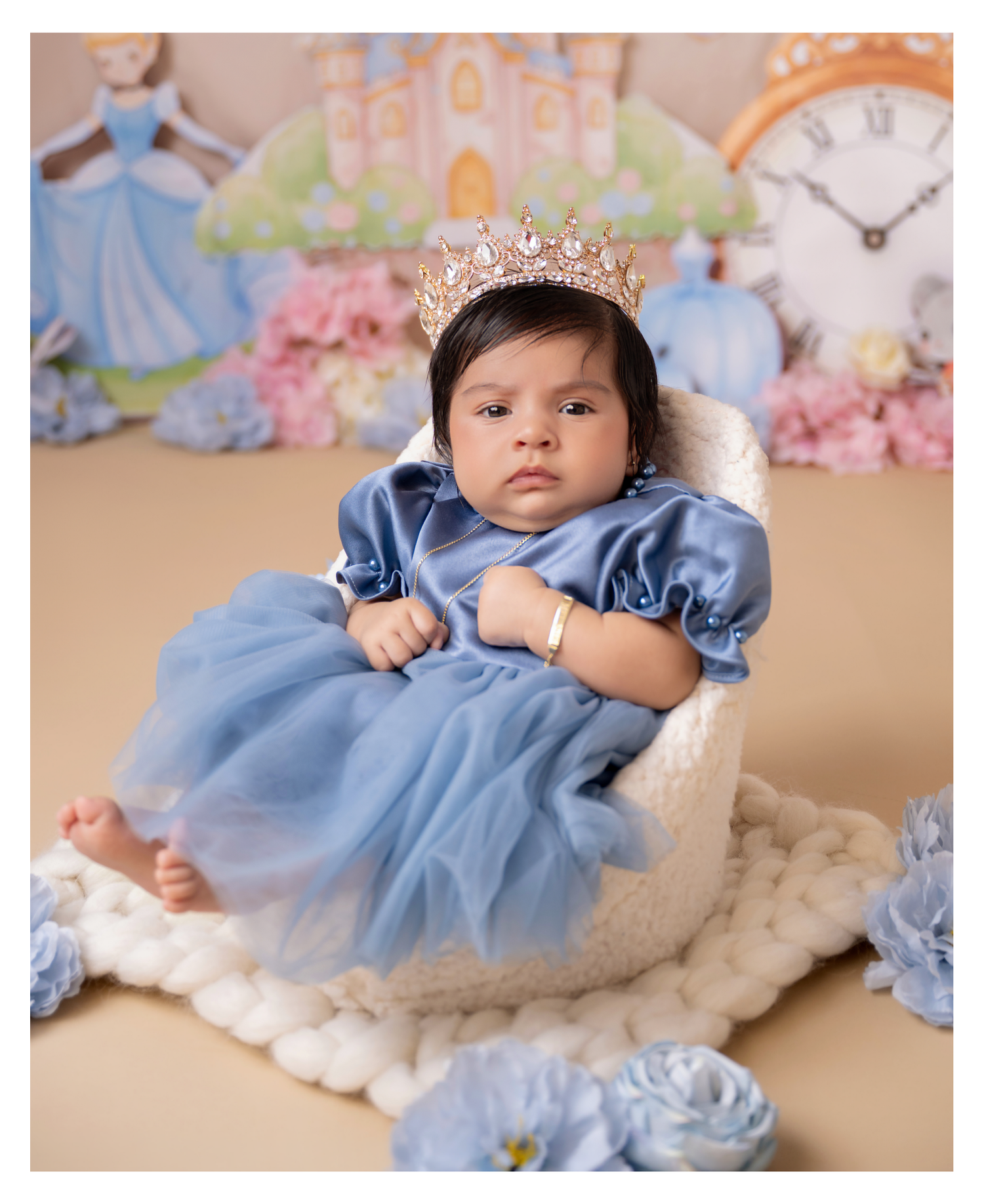 Baby girl in blue Cinderella dress and crown sits in white chair with princess castle backdrop.