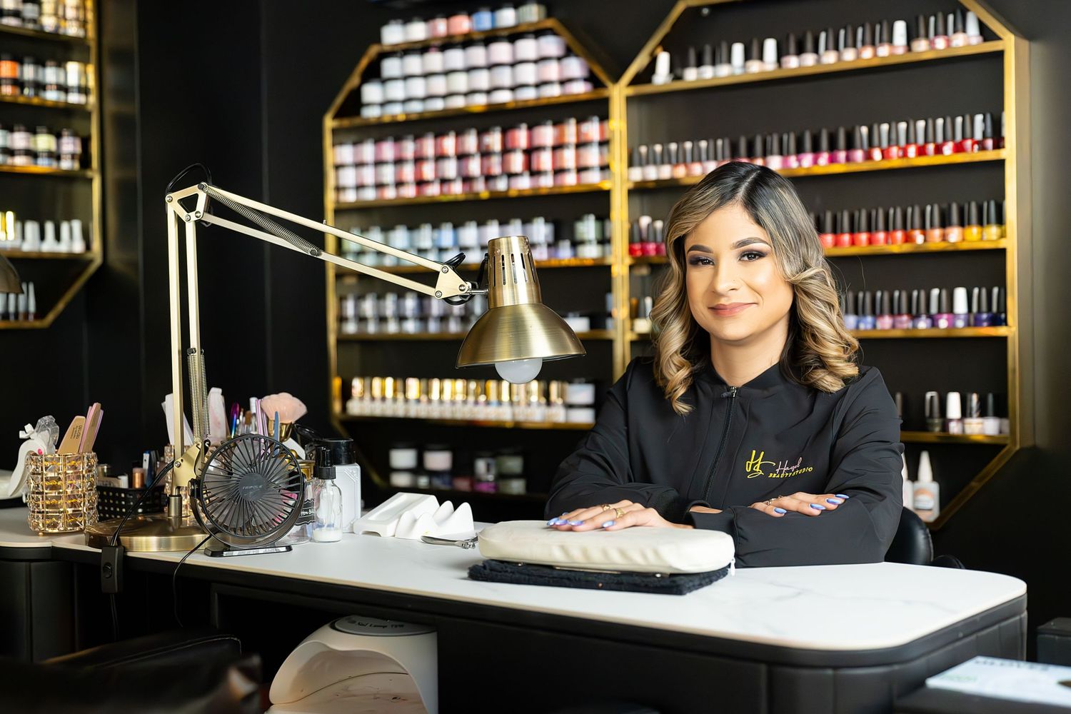 Nail technician poses at manicure station in modern salon with colorful nail polish display shelves in background.