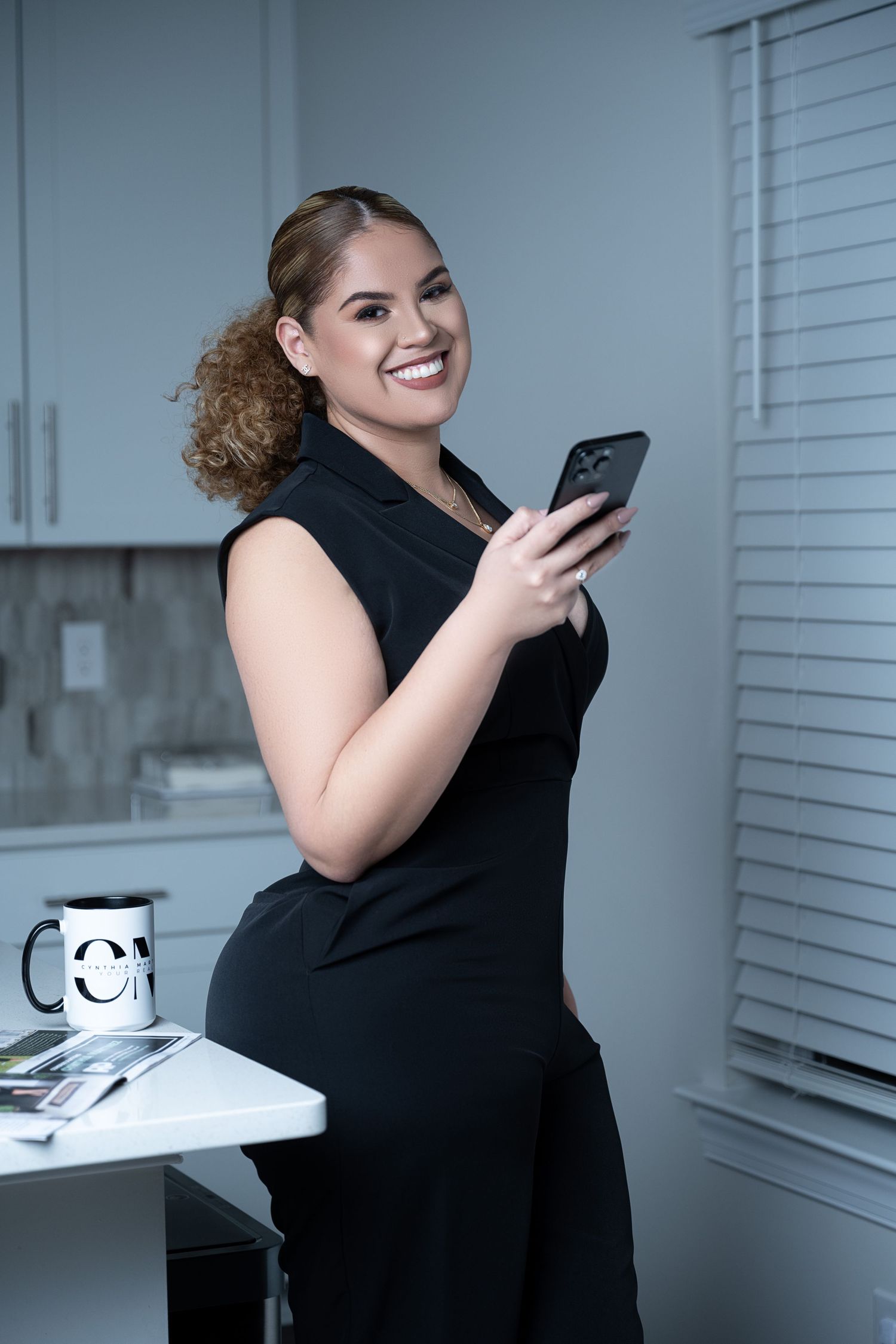 Smiling woman in black dress holding smartphone, leaning against white desk with coffee mug in modern office setting.