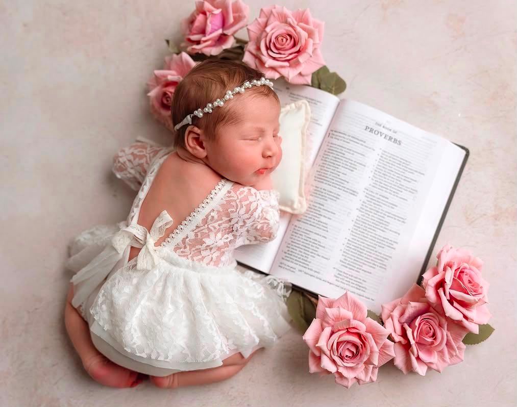 Sleeping newborn baby girl in white lace romper with pearl headband, surrounded by pink roses and an open Bible.