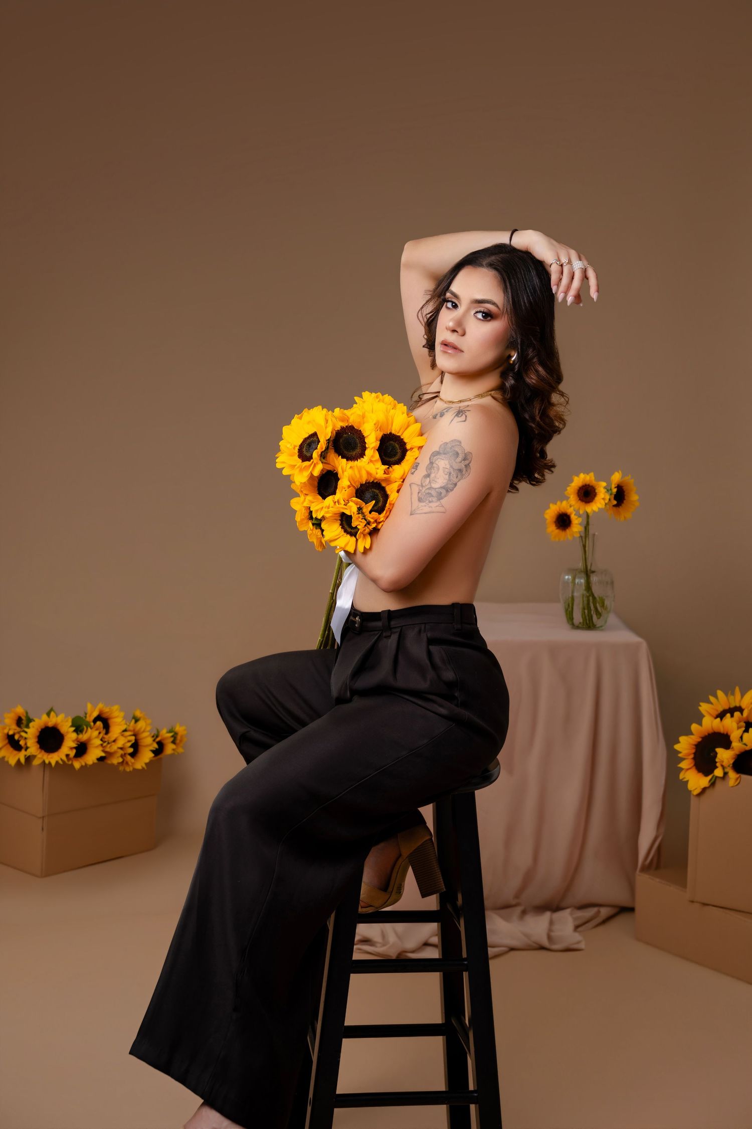 Woman with wavy dark hair sits on a black stool holding a sunflower bouquet in black trousers against a tan studio backdrop.