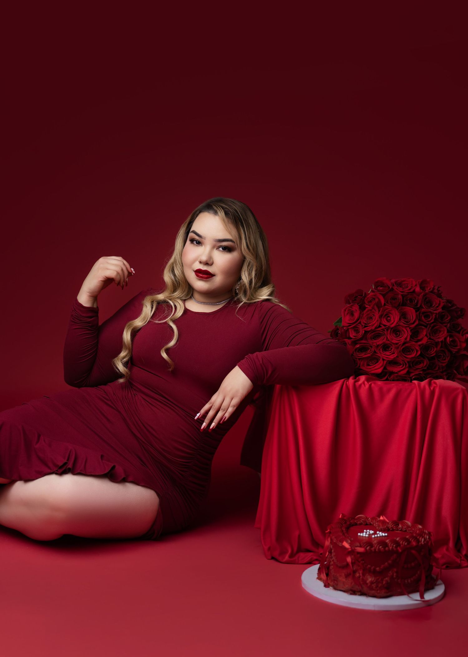 Woman in a burgundy dress with wavy blonde hair poses on a red backdrop beside a red rose bouquet and a rose-decorated cake.
