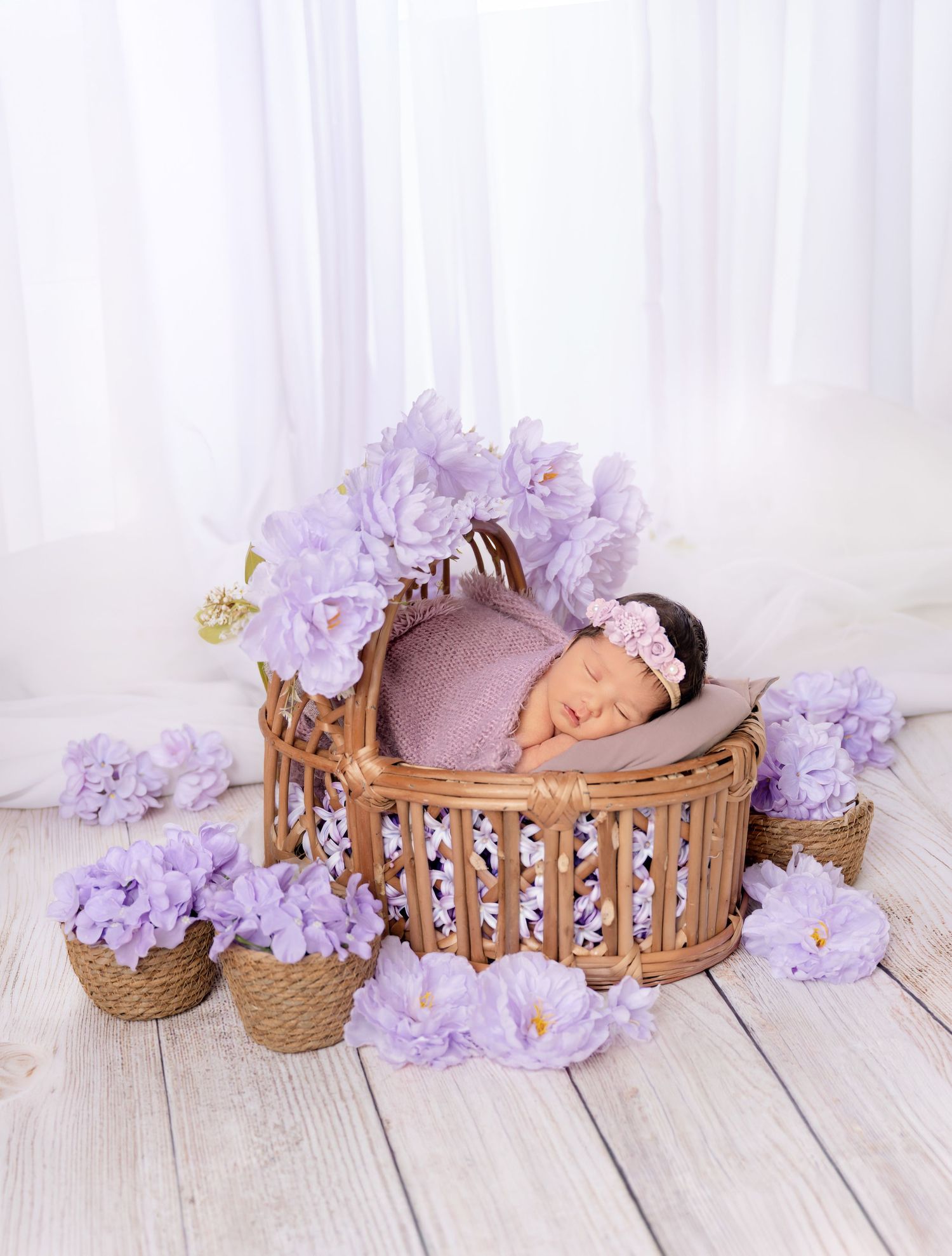 Newborn baby sleeping in wicker basket surrounded by purple lavender flowers with floral headband on white wood floor.