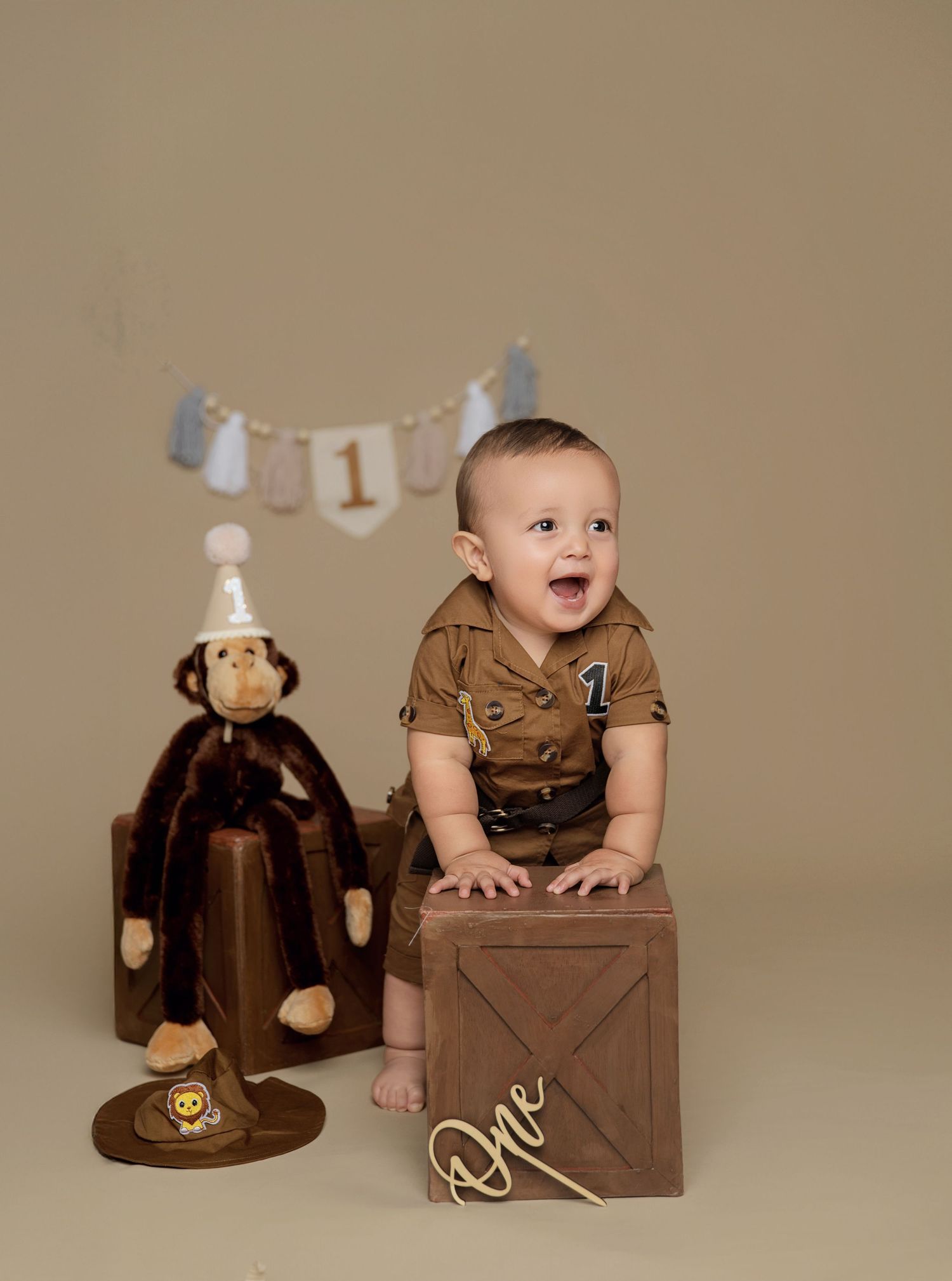 Baby's first birthday photo with a monkey stuffed animal, wooden crate, and number one banner on beige background.