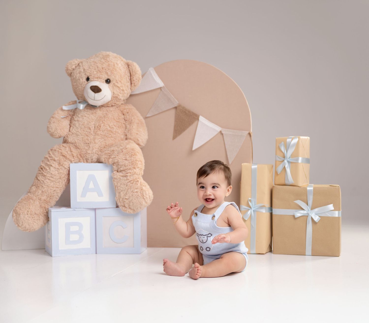 Baby boy sitting beside a large teddy bear on ABC blocks and wrapped gifts in a soft neutral studio setting.