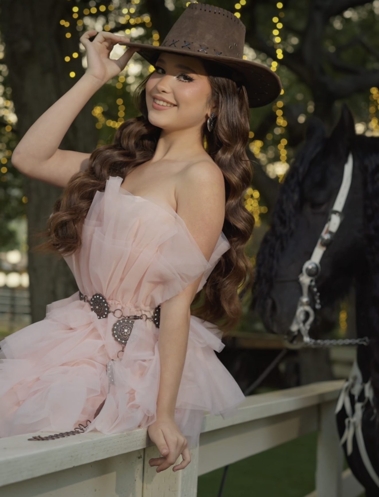 Young woman in pink tulle dress and brown cowboy hat poses by a fence with a black horse outdoors.