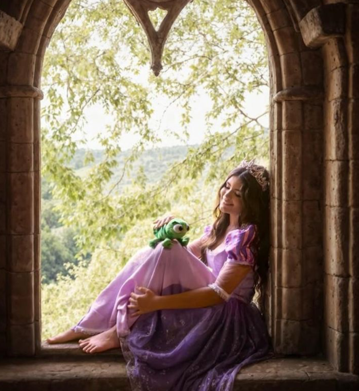 A young woman in a purple Rapunzel gown sits in a stone arch window holding a frog plush toy.