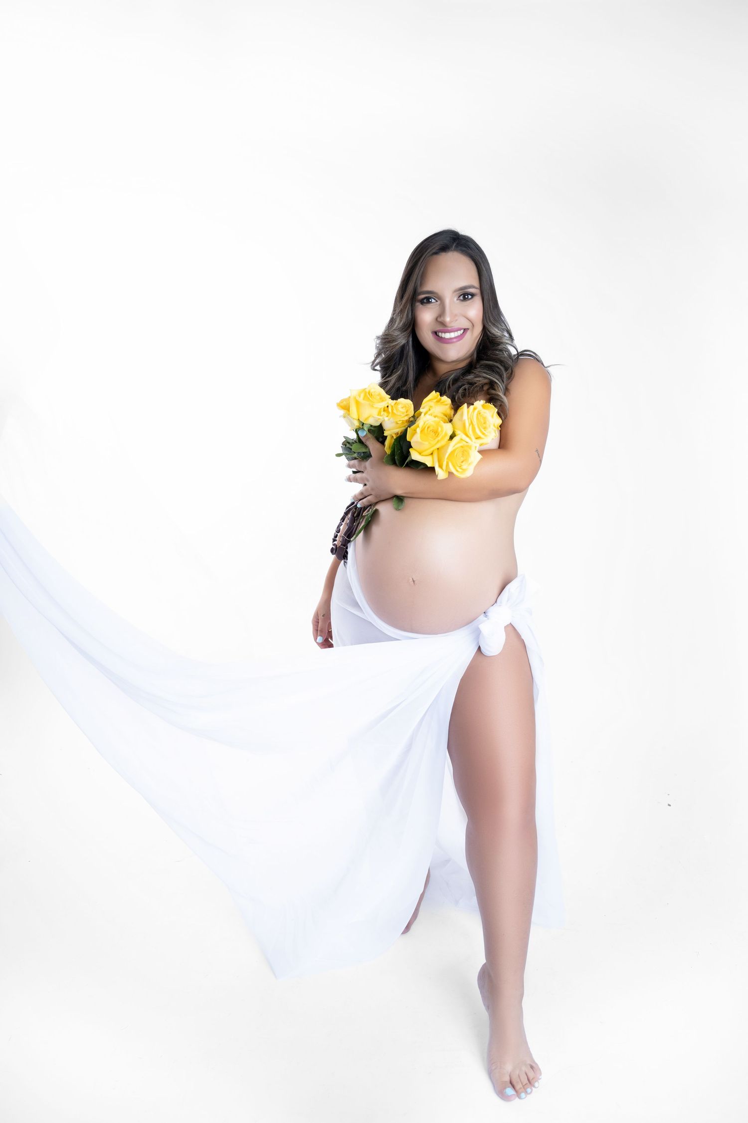 Pregnant woman holding yellow roses bouquet, wrapped in white flowing fabric, smiling against white background.