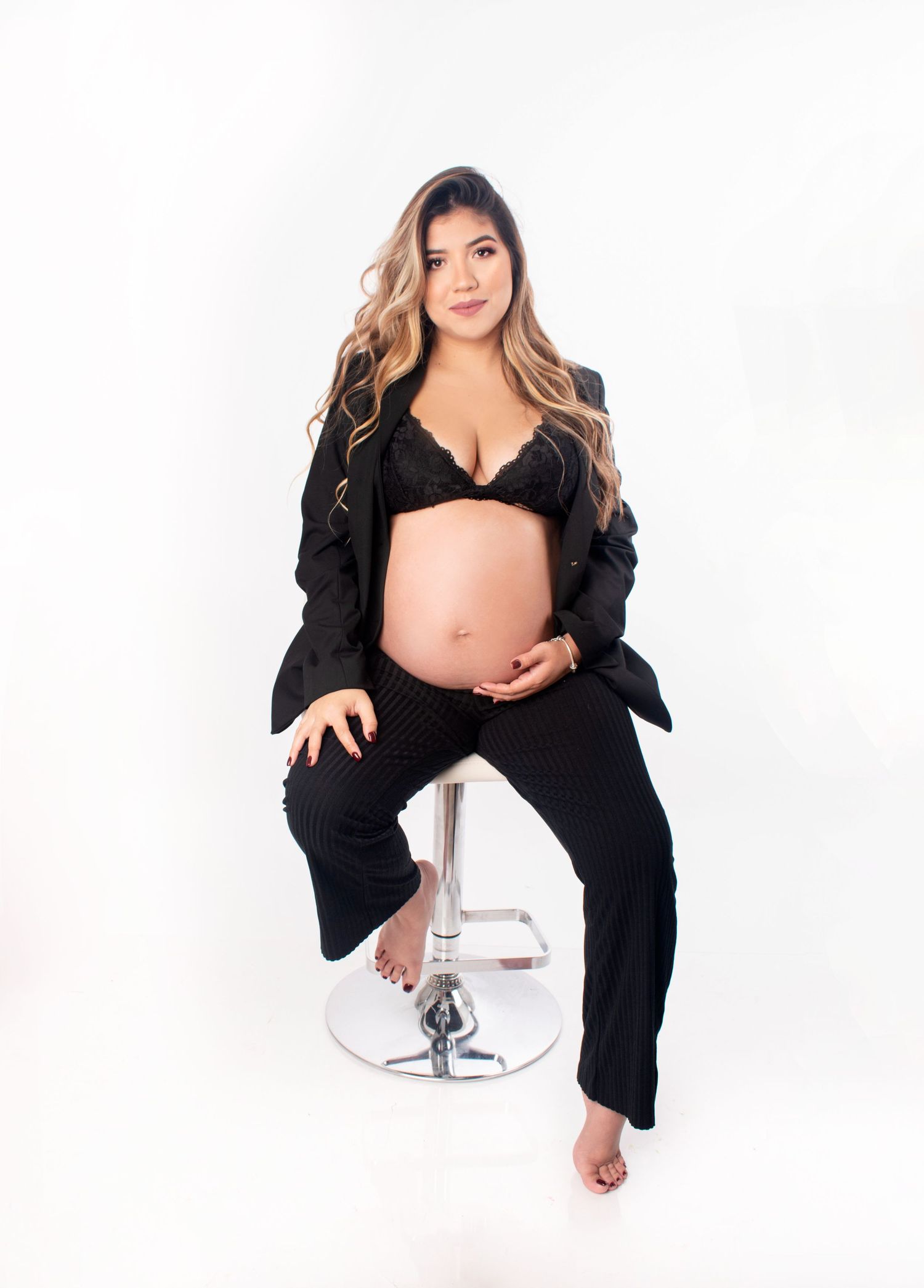 Pregnant woman in black lace bralette and blazer sitting on stool, showcasing baby bump in studio maternity photo.