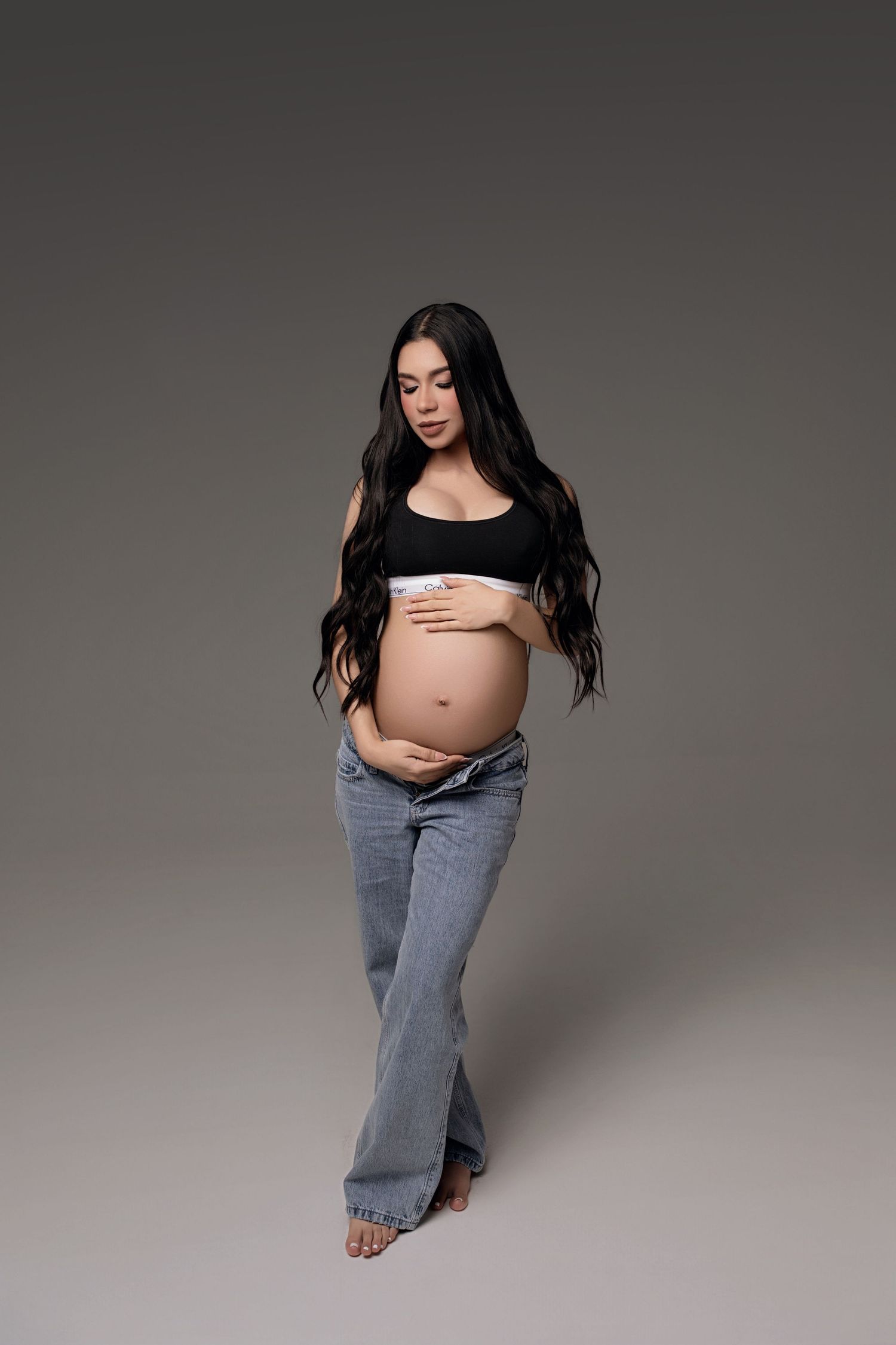 Pregnant woman in black crop top and gray flare jeans cradling baby bump, posing on gray background.