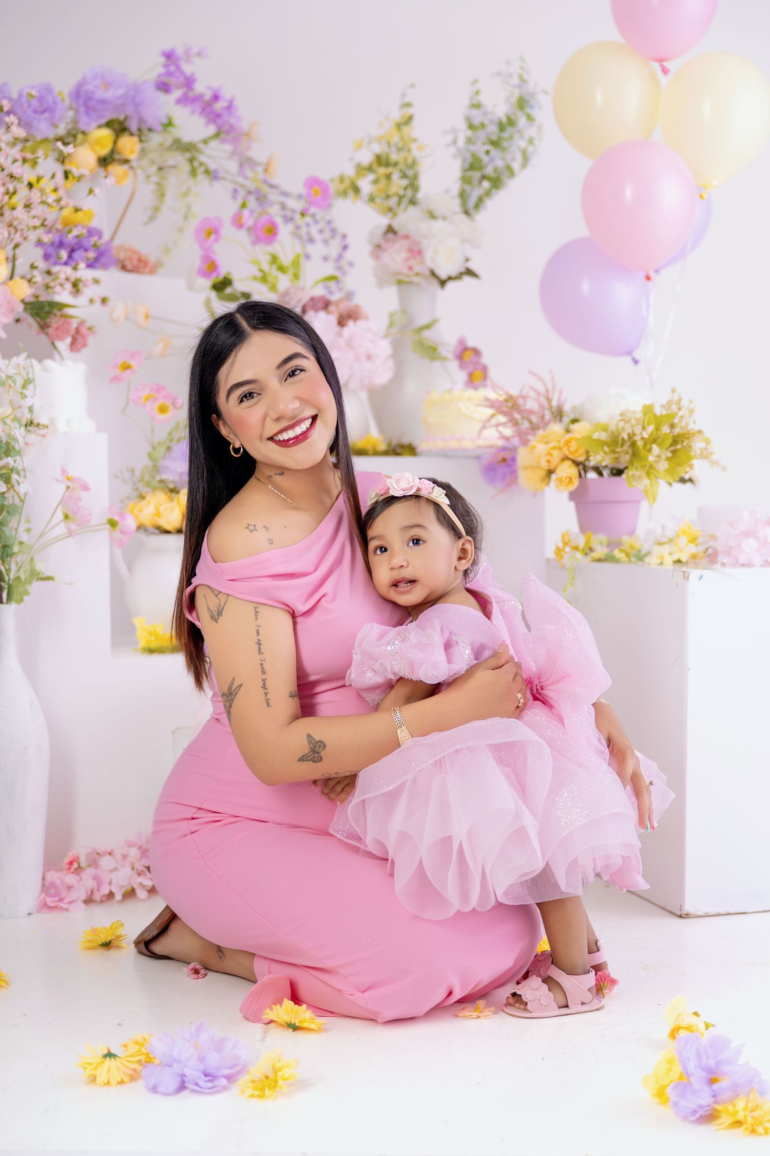 A smiling woman in a pink dress holds a baby girl in a pink tutu, surrounded by colorful flowers and balloons.