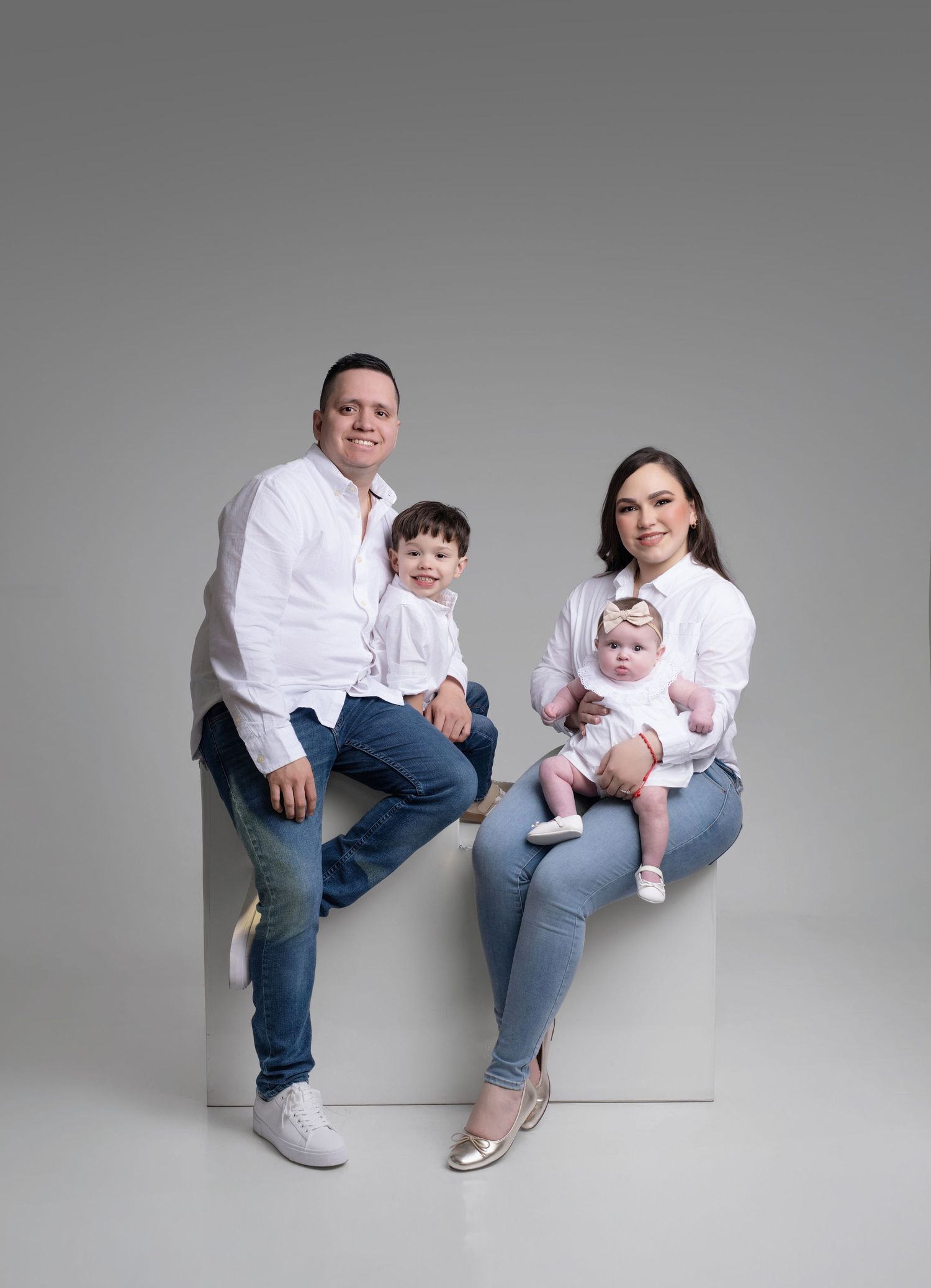 Family of four in white shirts and jeans posing for a studio portrait with two young children on a white cube.