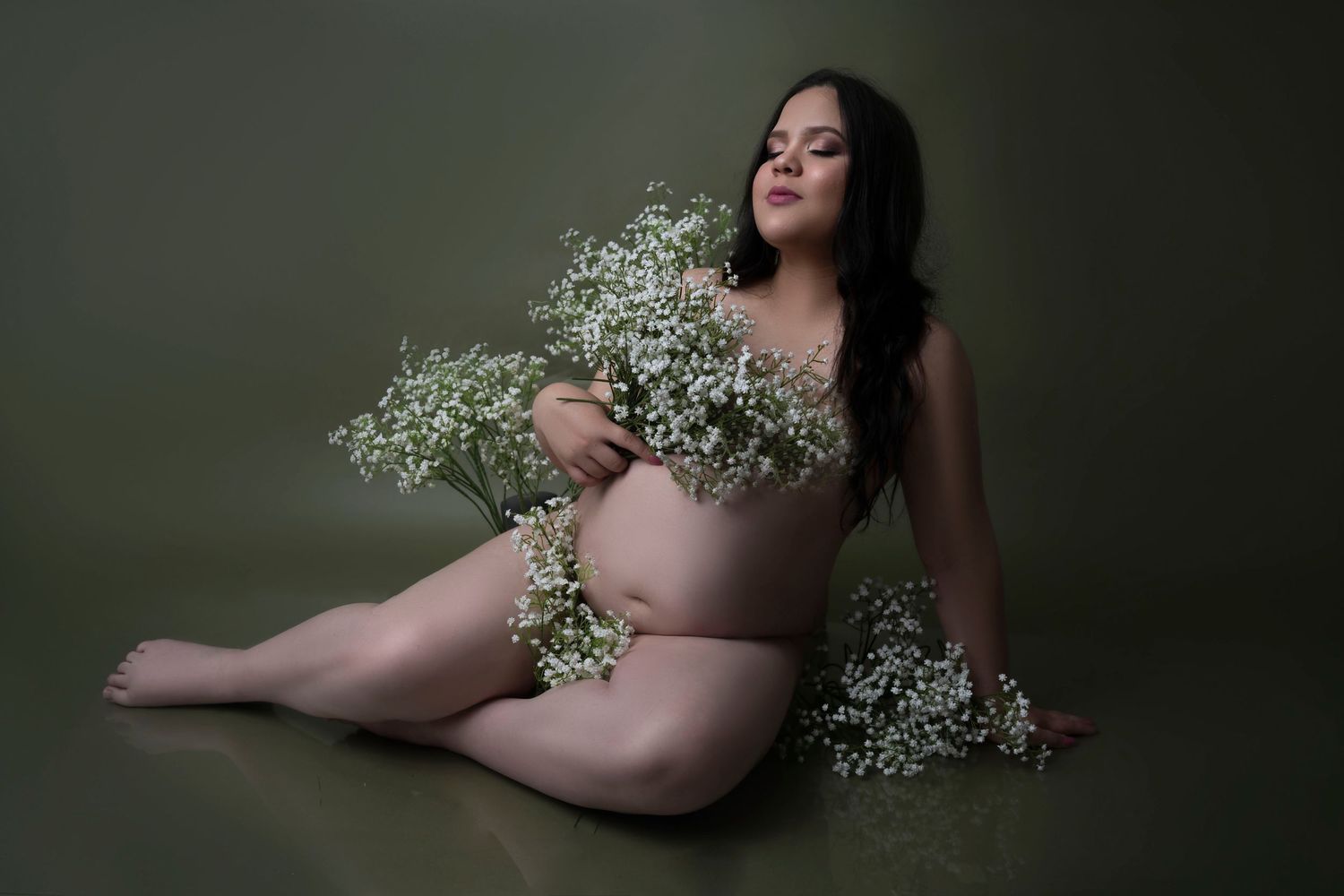 Pregnant woman sitting gracefully with white baby's breath flowers against an olive green background.