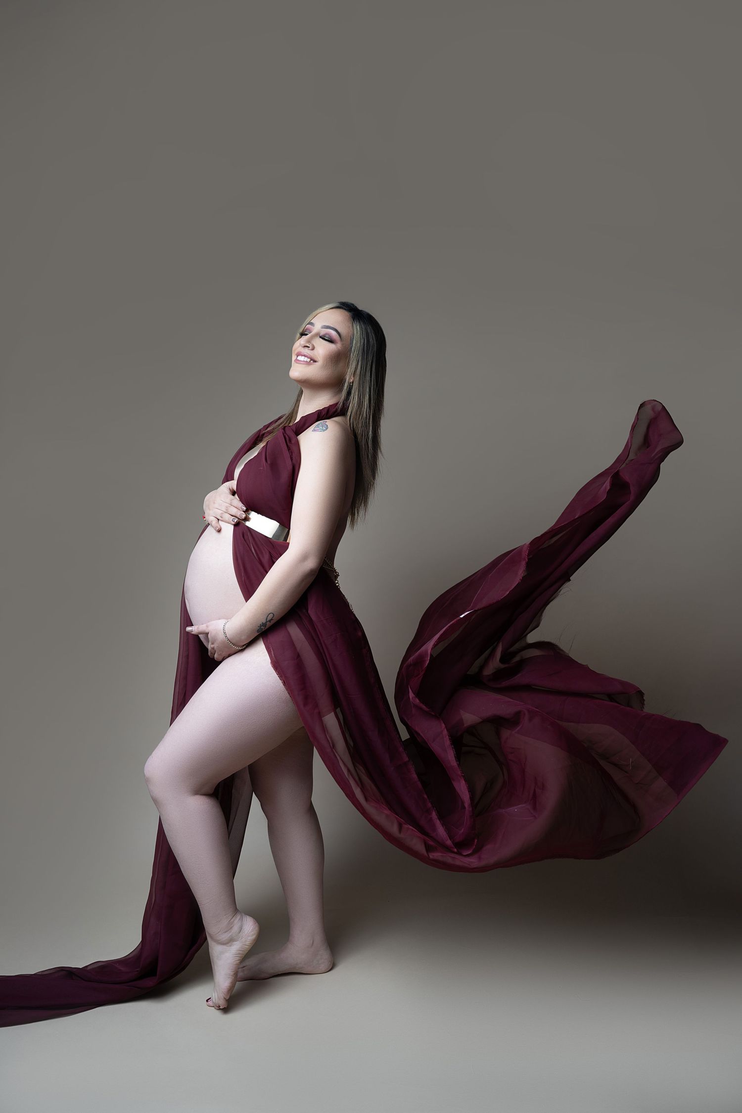 Smiling pregnant woman in flowing burgundy fabric draped elegantly, cradling her baby bump in a studio setting.