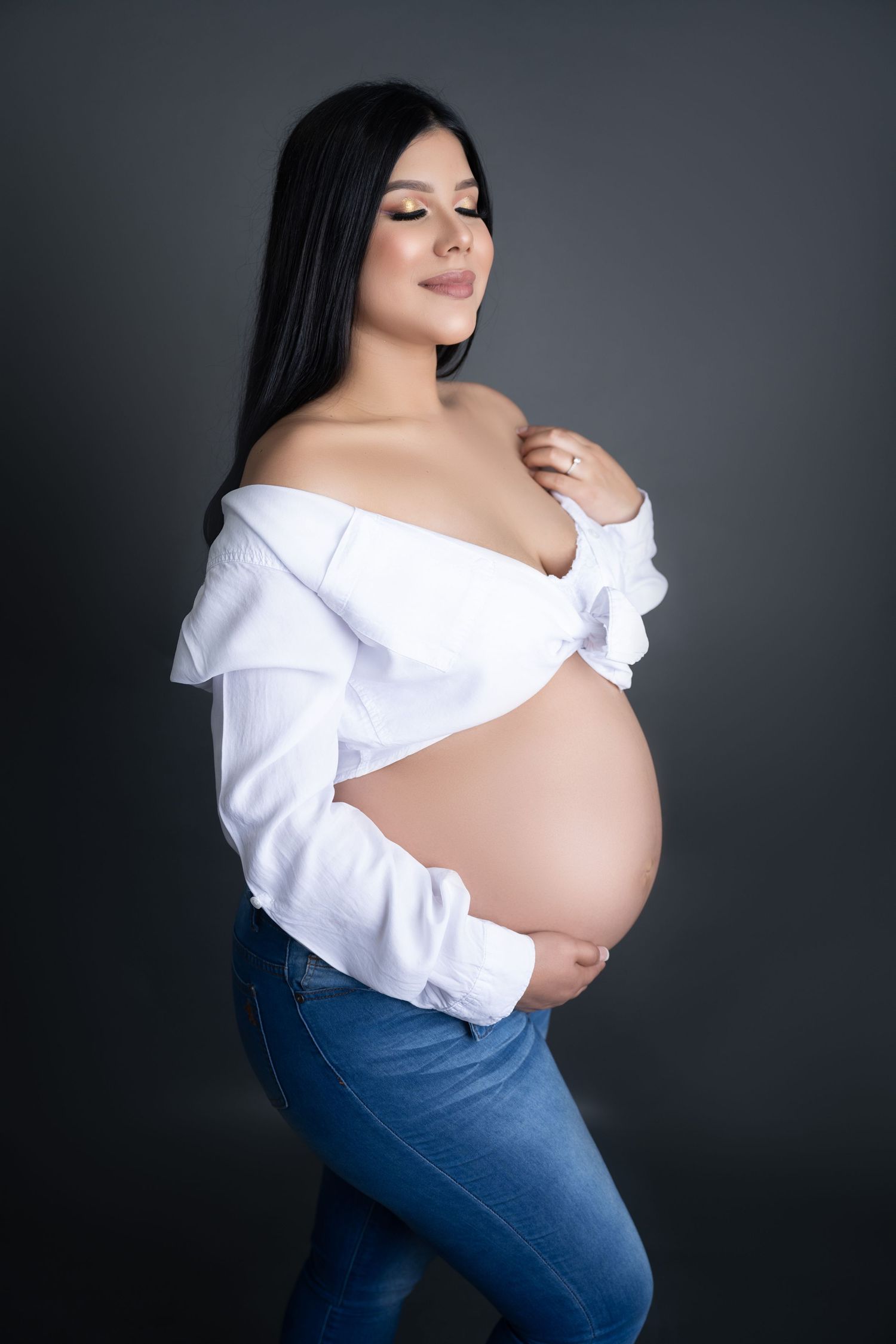 Pregnant woman in white off-shoulder top and jeans cradling baby bump, studio maternity portrait on dark background.