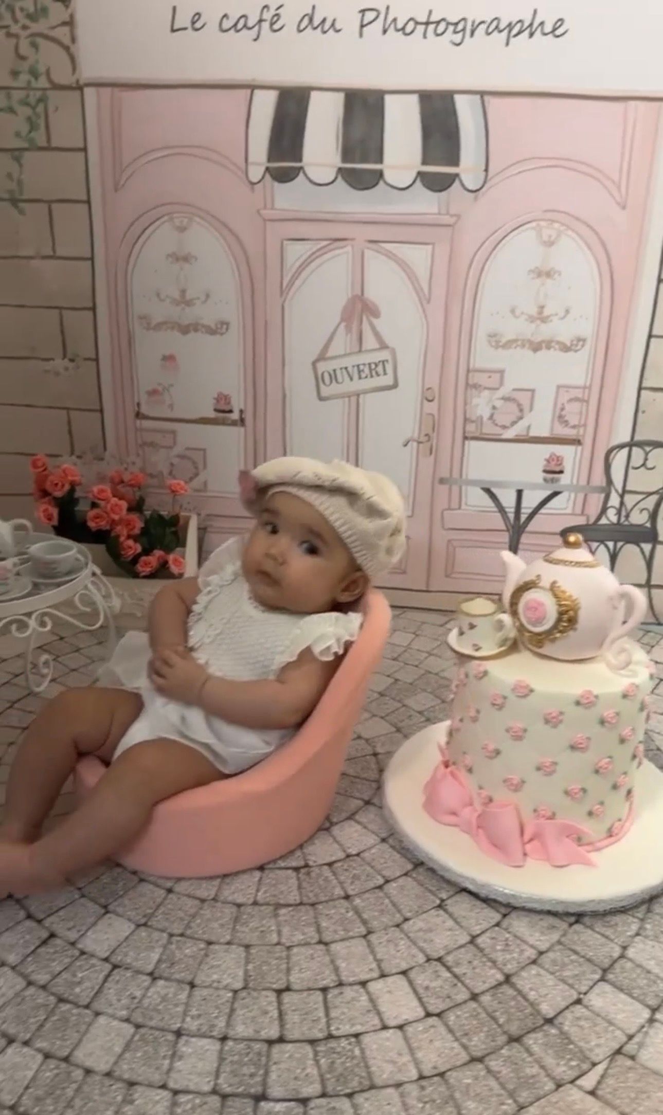 Baby girl in white outfit and beret sits beside a floral tea party cake at a French cafe themed photo shoot.
