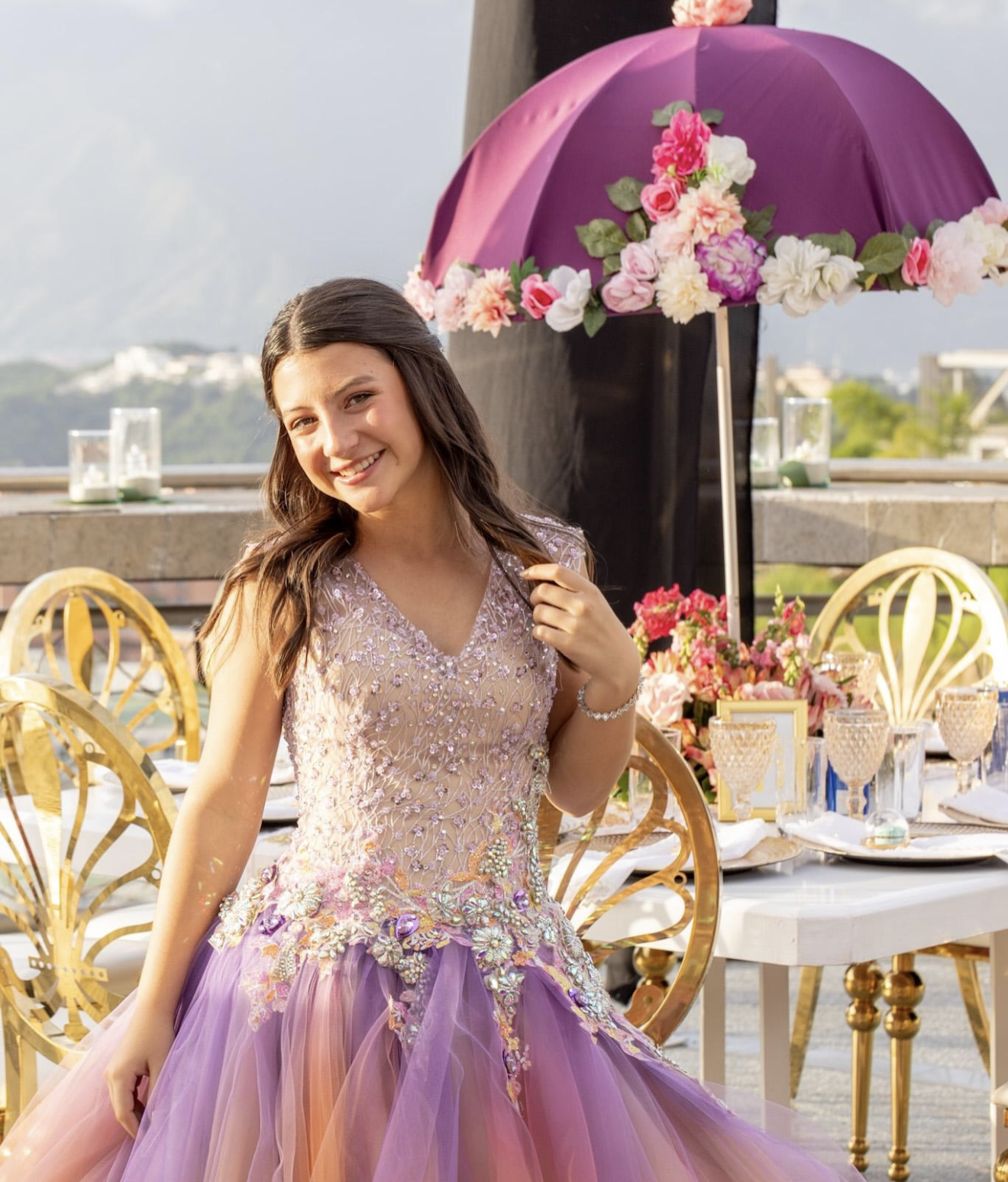 Young woman in lavender floral ballgown quinceañera dress poses outdoors near decorated table with purple floral umbrella.