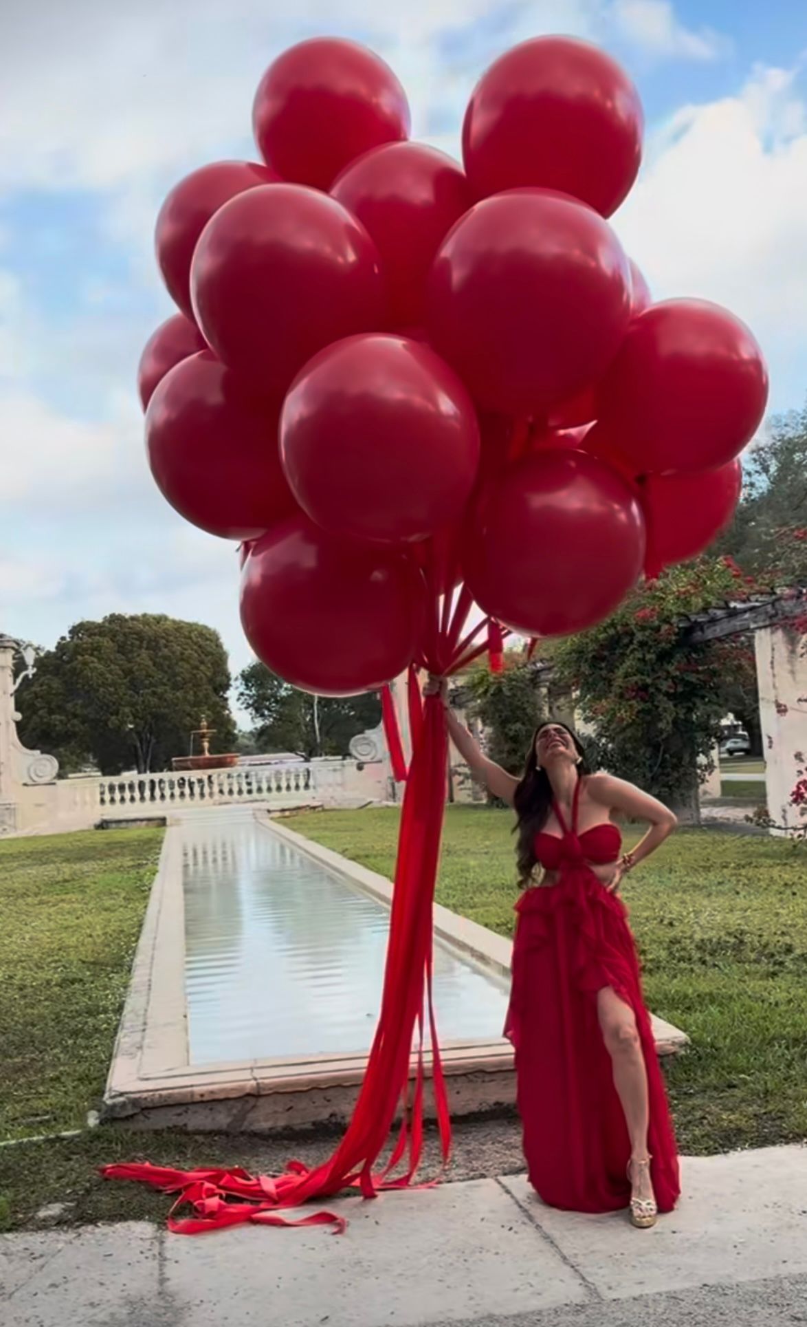 Woman in red gown holding bundle of red balloons beside reflecting pool in elegant garden setting.