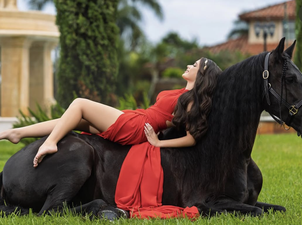 Woman in red dress reclining on a black horse lying in grass near a mansion.
