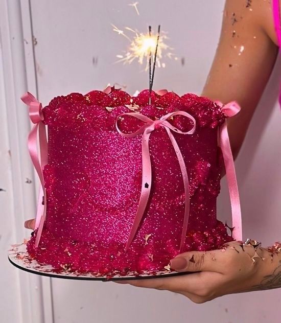 A dazzling hot pink glitter cake with ribbon bows and a sparkler on top, held by a person at a birthday celebration.