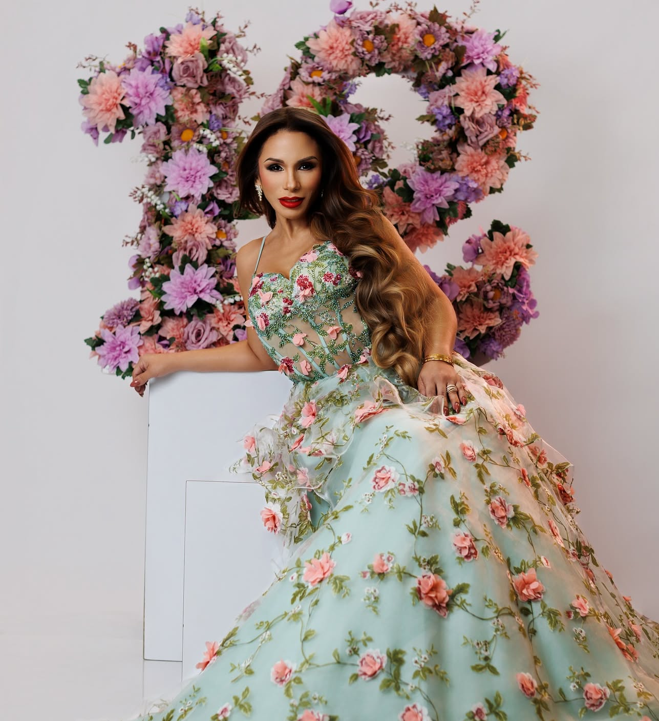 Woman in floral ball gown poses elegantly against a heart-shaped flower wall backdrop.