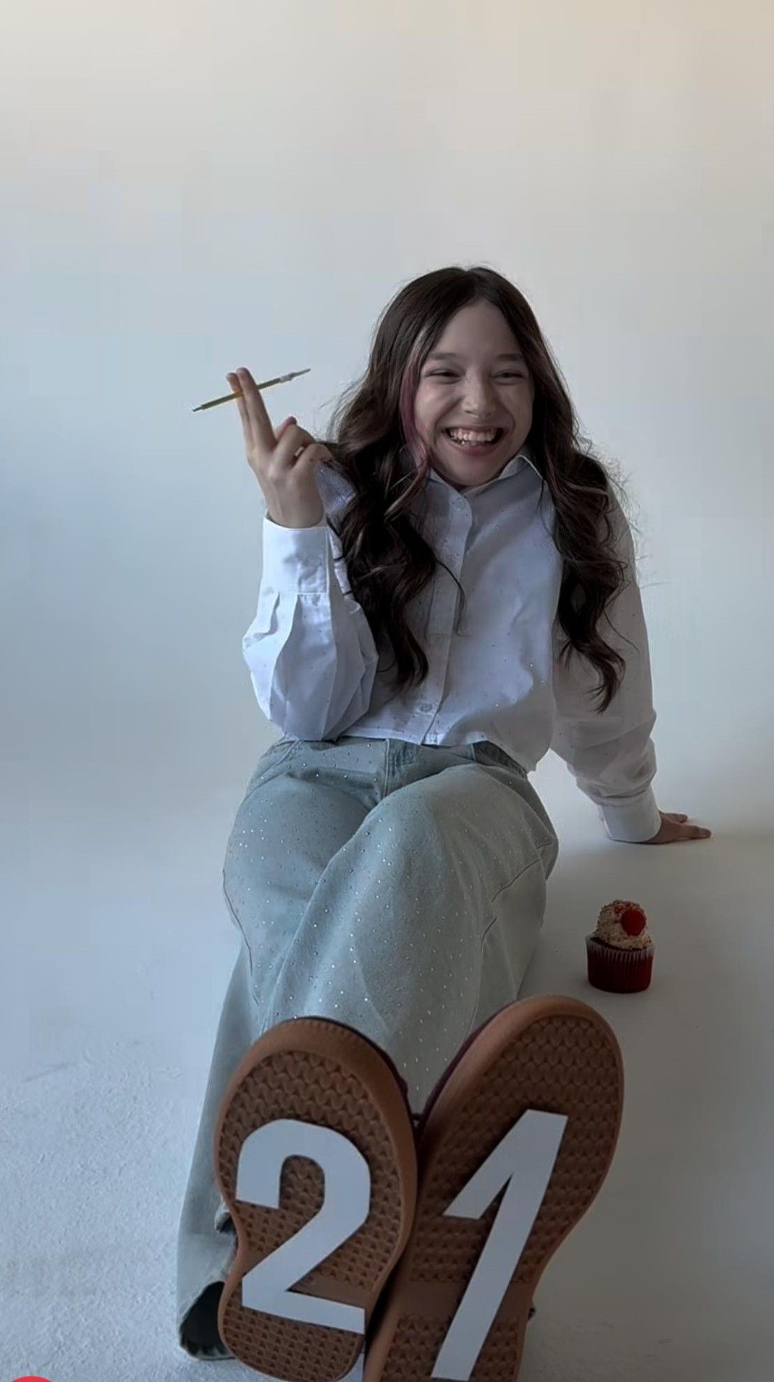 Young woman in white shirt and mint skirt sitting, making rock sign gesture, shoes showing number 21.