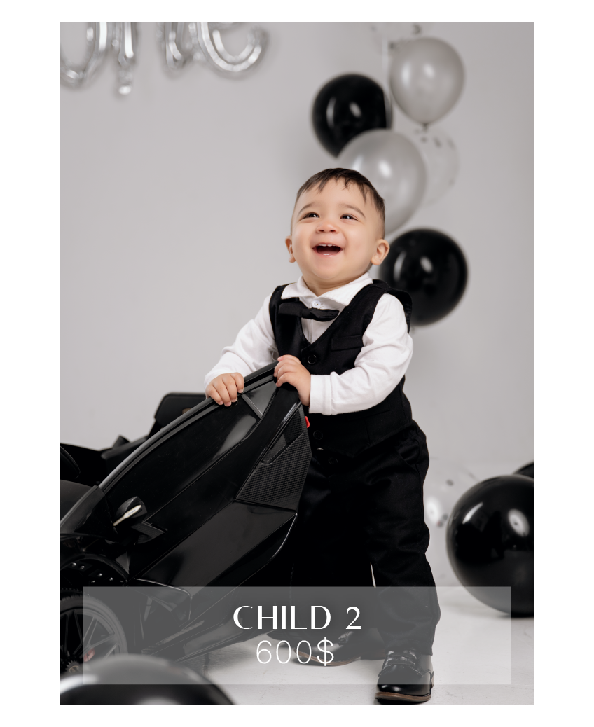 Smiling baby boy in black tuxedo outfit posing with toy motorcycle surrounded by black and silver balloons.