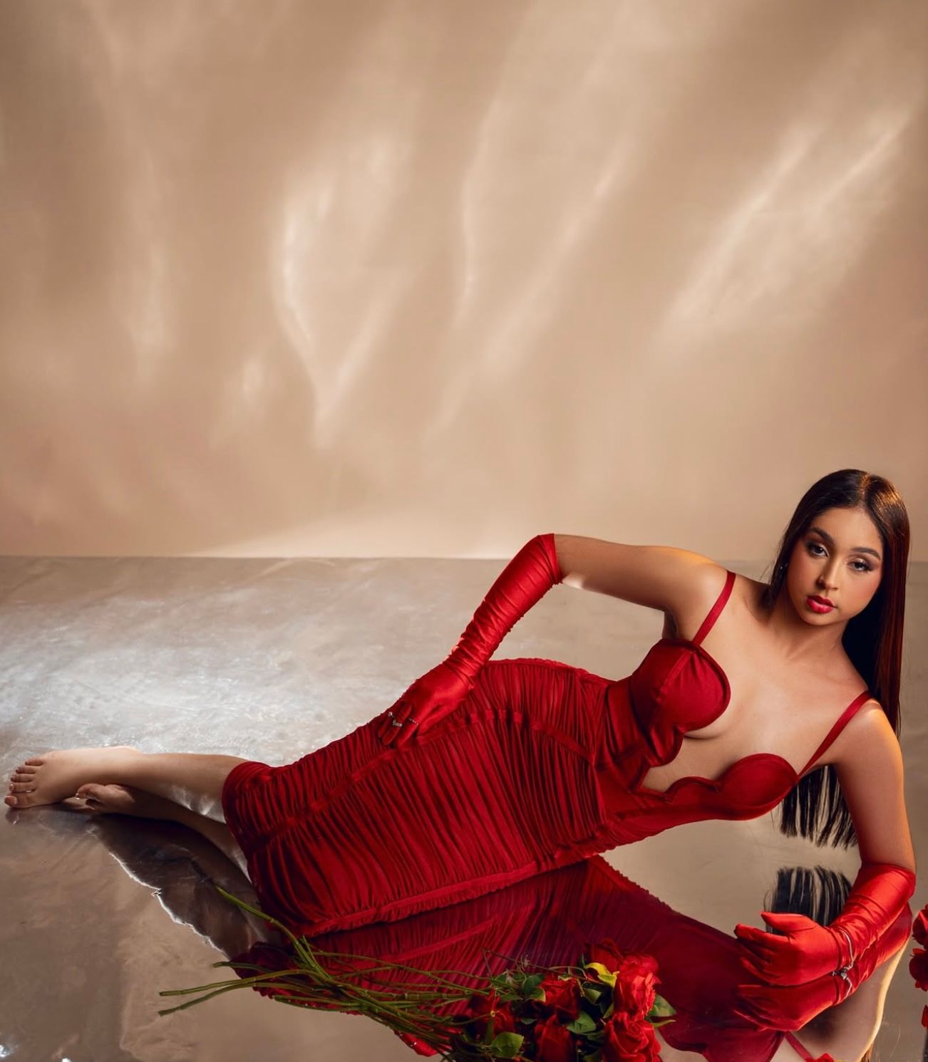 Woman in red ruched dress and long gloves reclines elegantly on floor with red roses nearby.