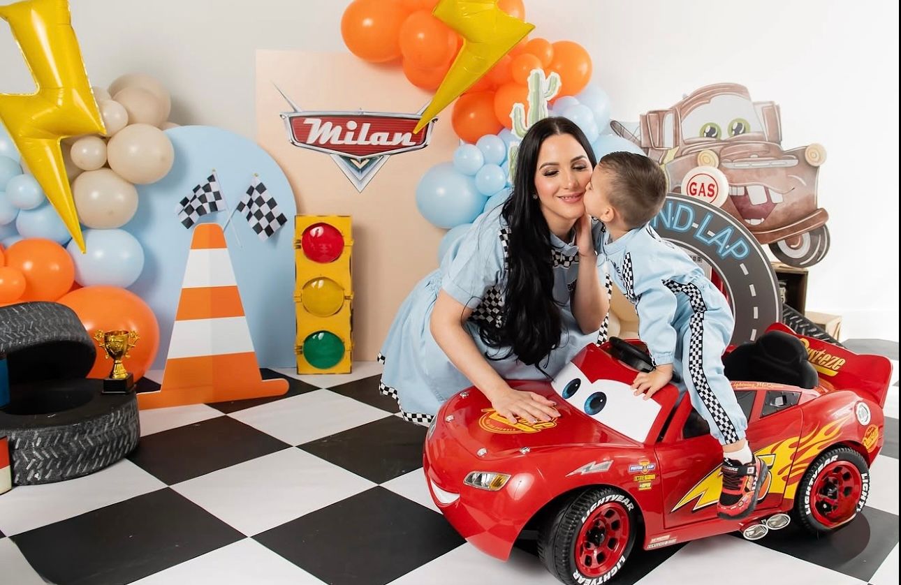 Race car themed party setup with checkered floor, traffic cones, balloons, and a red toy car against party decorations.