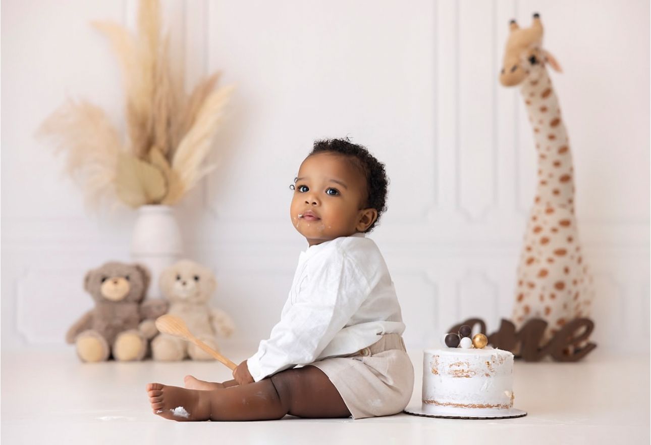 Baby in white outfit sits on floor surrounded by plush toys and giraffe decor for first birthday photo shoot.