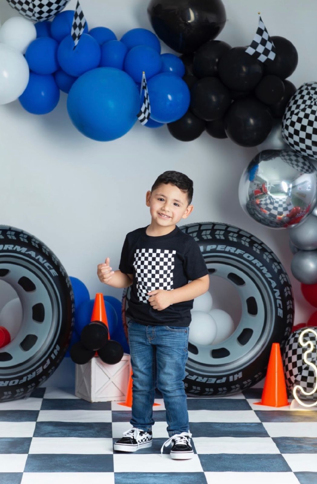 Racing-themed birthday setup with checkered flags, blue and black balloons, and orange traffic cones on a checkered floor.
